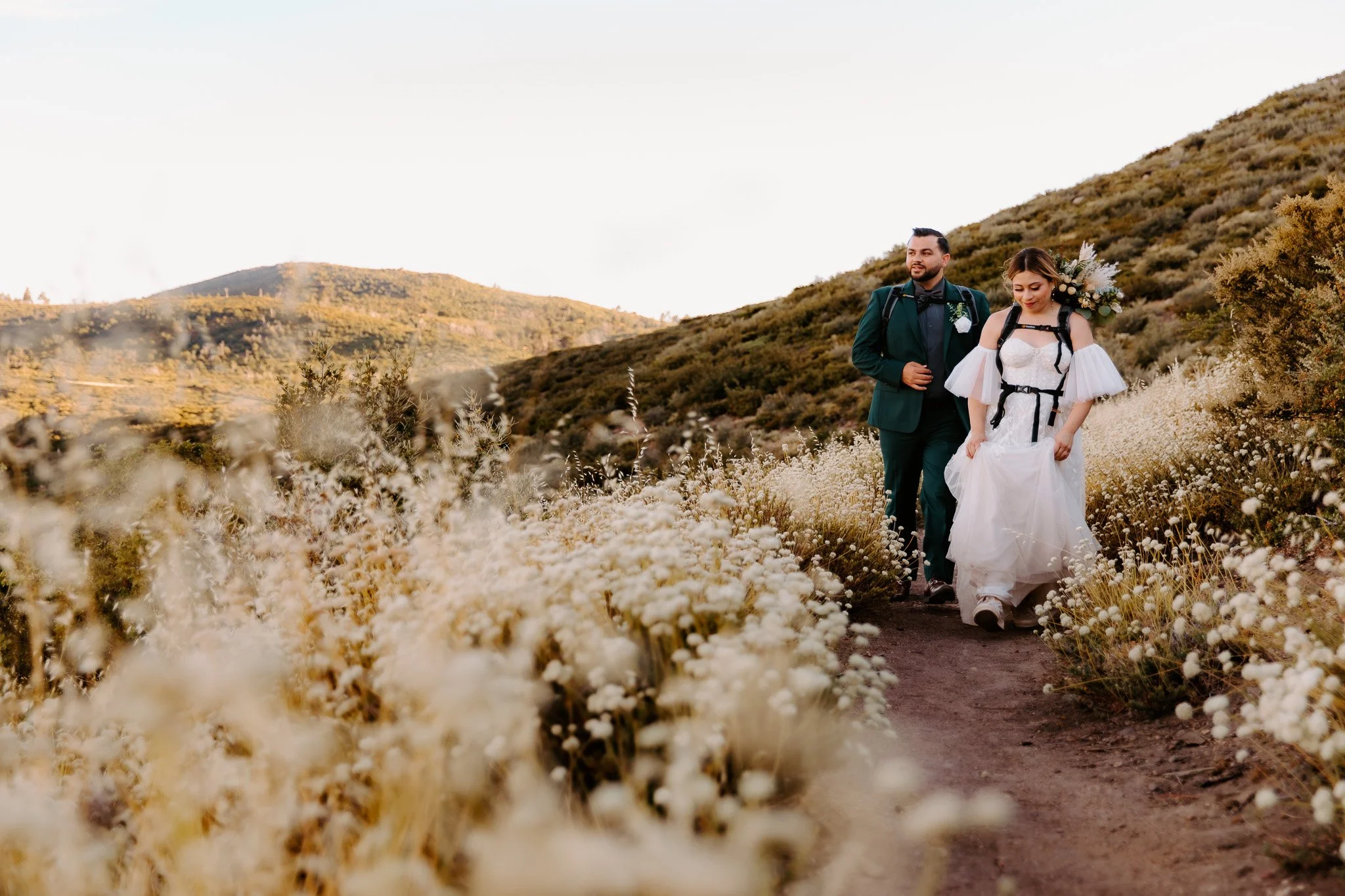 A bride and groom walking through a field of white flowers with mountains in the background, during what appears to be sunset.
