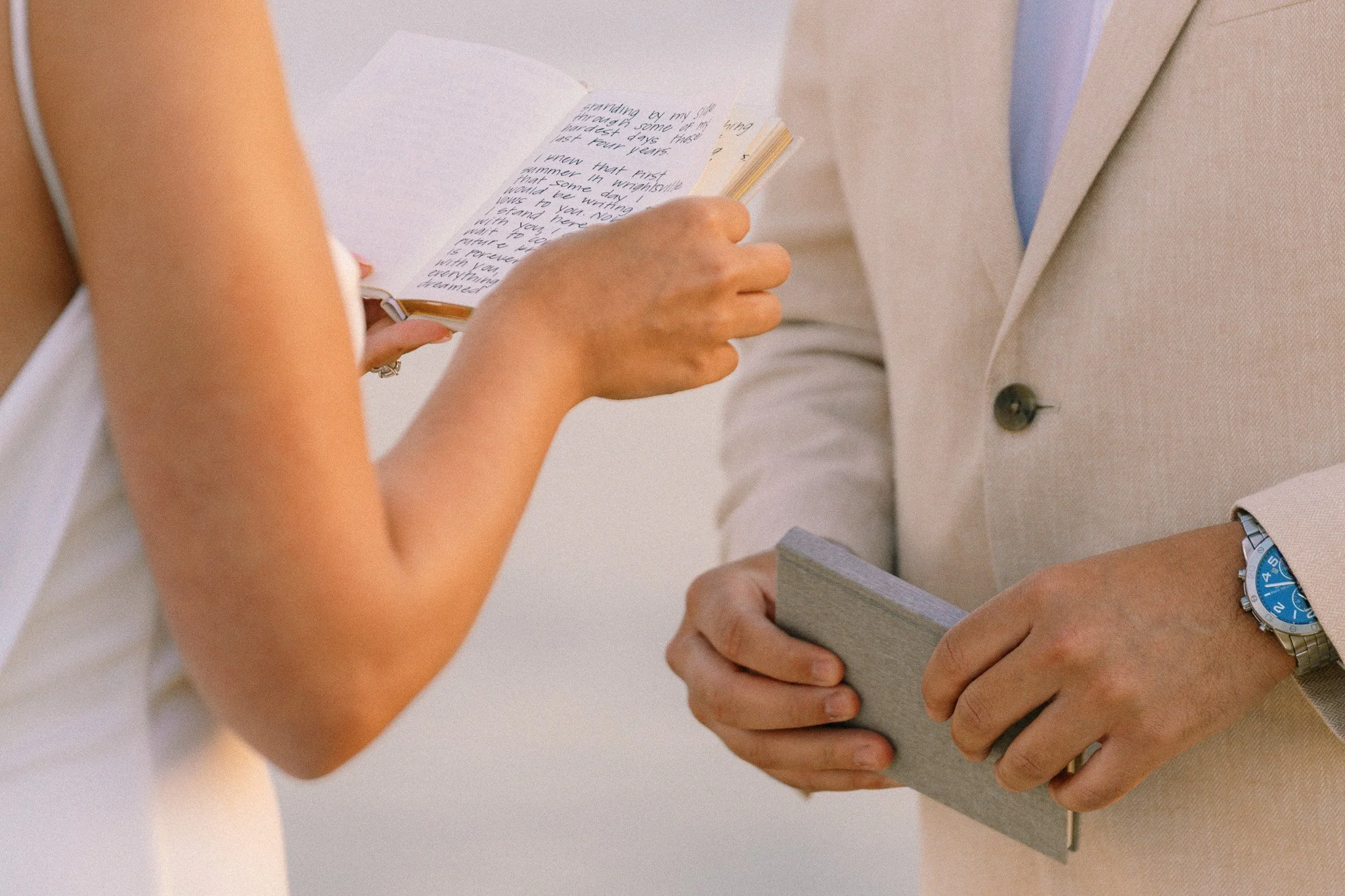 Close-up of two people exchanging gifts. One person is holding a small book with cursive writing, the other is holding a stack of books, both dressed in light-colored clothing, with a white background.
