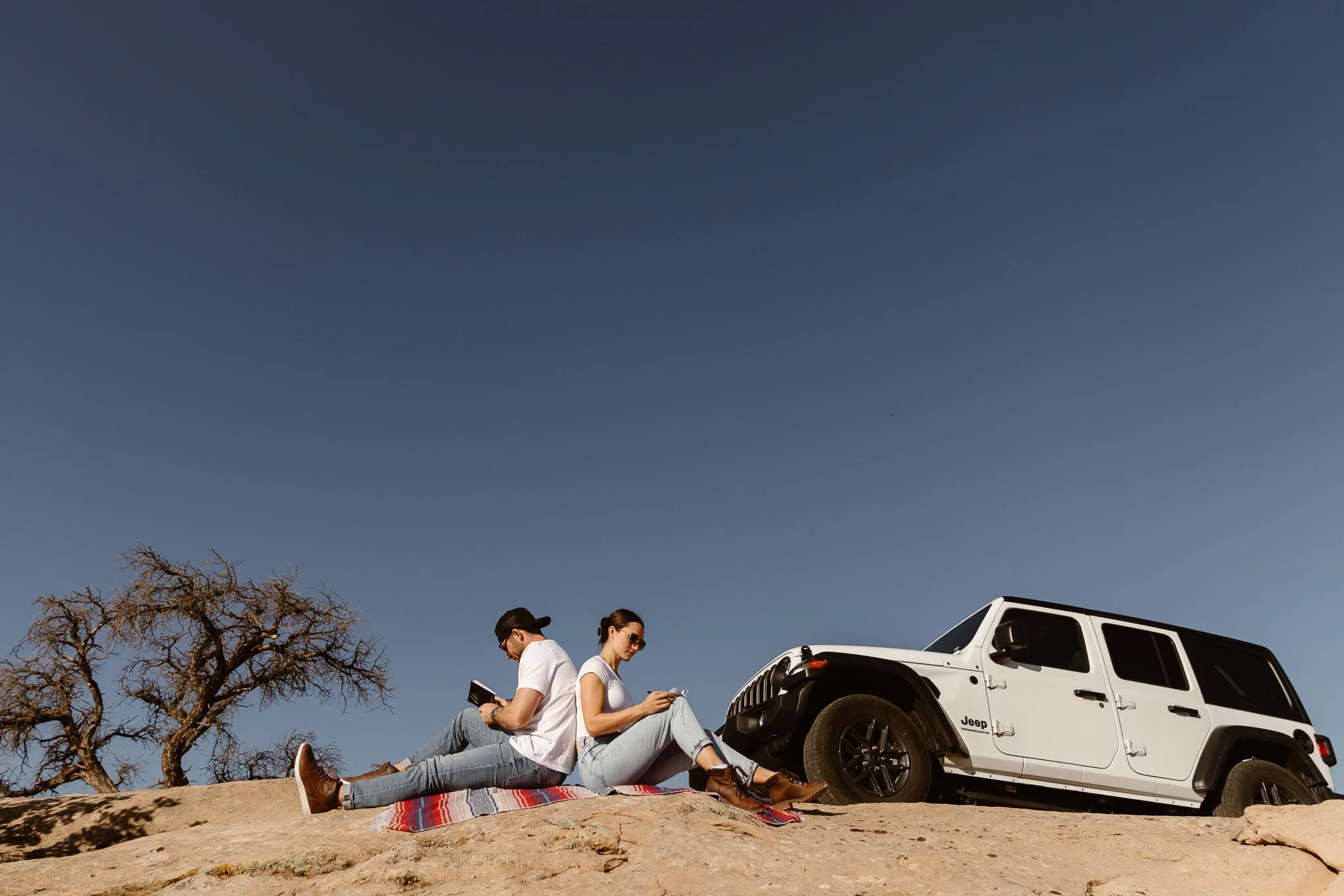 Two people sitting on a blanket on rocky ground near a white Jeep parked beside them, under a clear sky with a bare tree in the background.