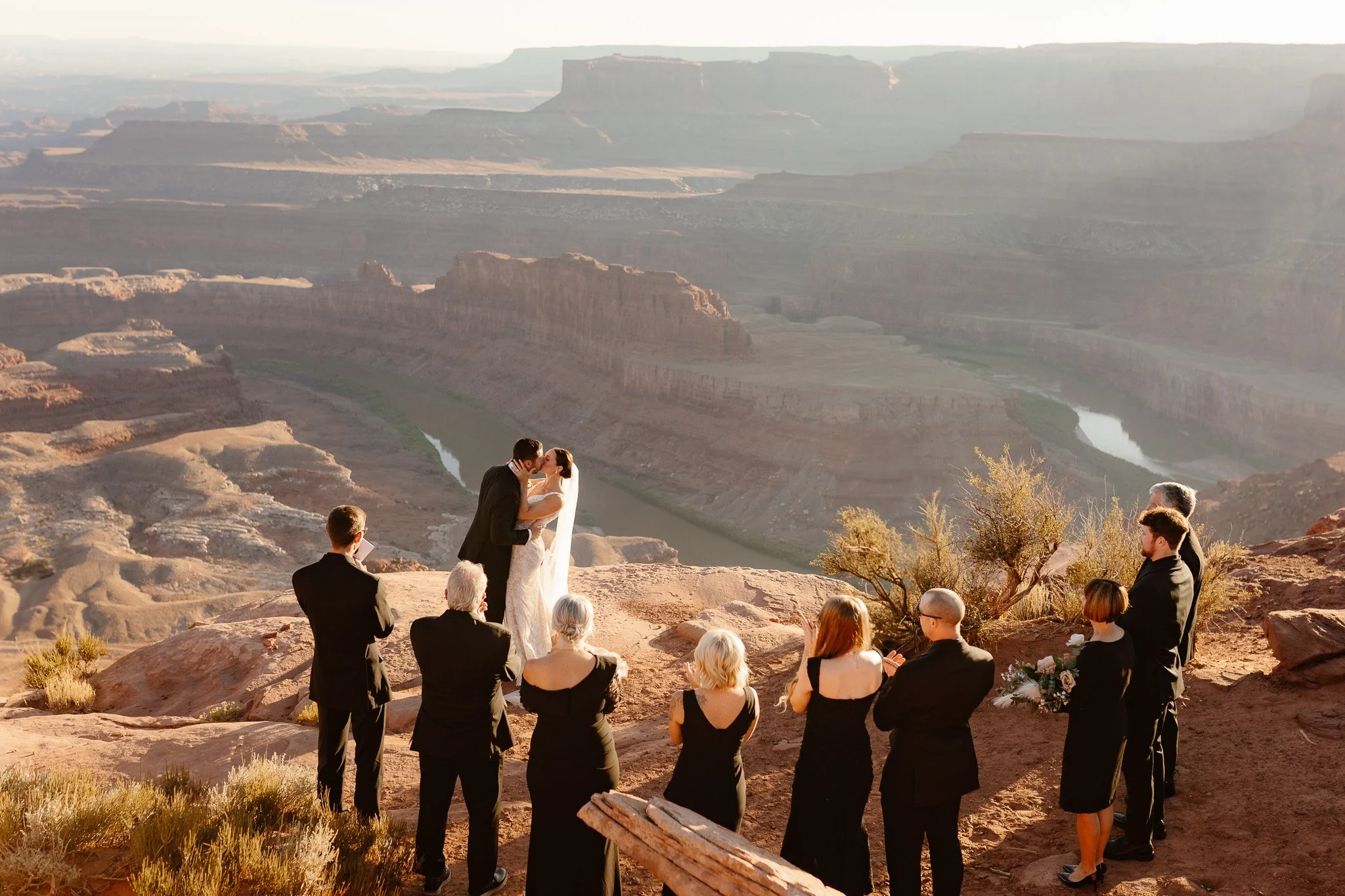 A wedding ceremony at the Grand Canyon during sunset, with the bride and groom kissing as the officiant presides and guests watch.