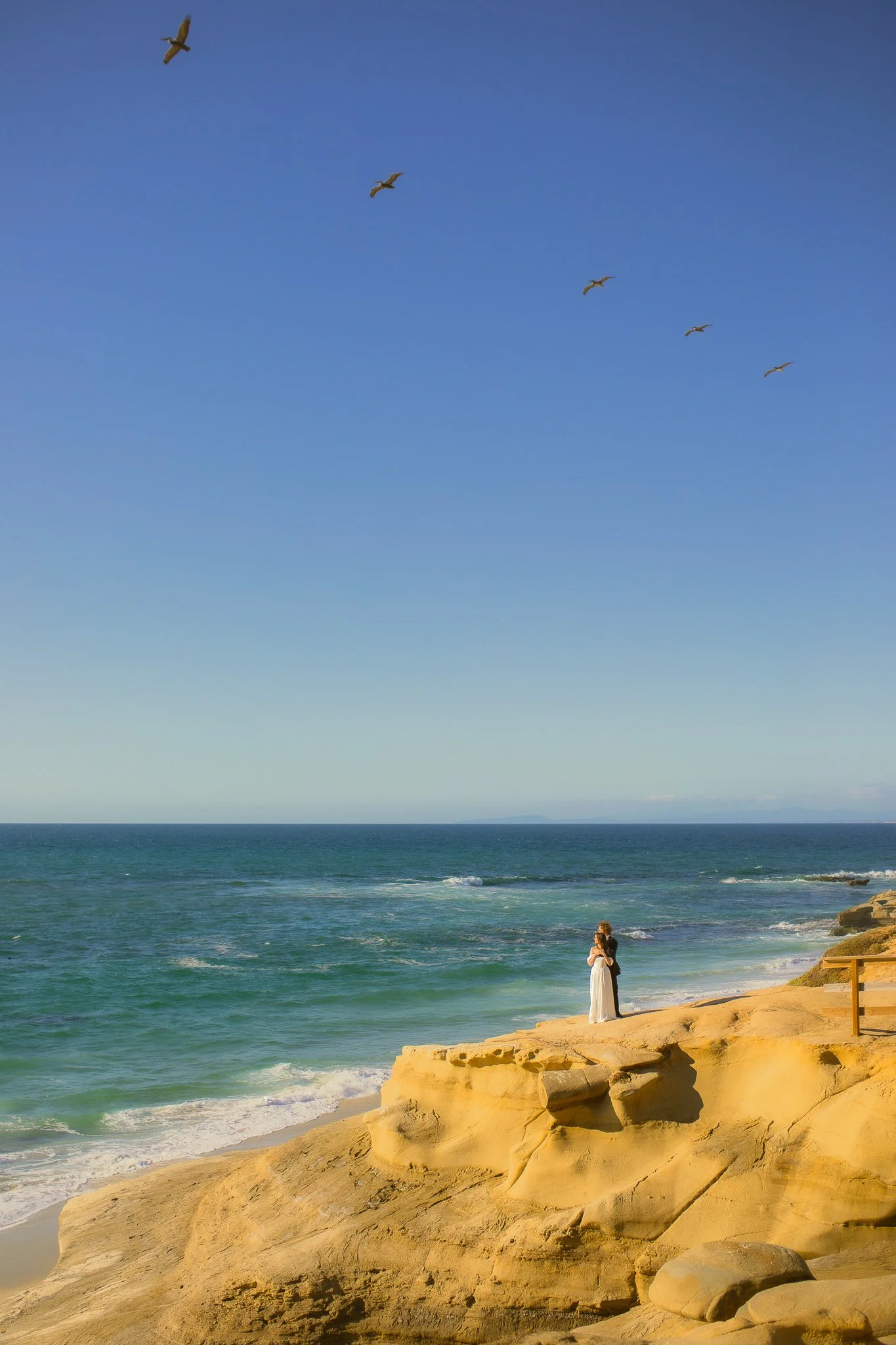 A woman in a white dress standing on rocks at the beach, with the ocean and a blue sky behind her and four seagulls flying overhead.