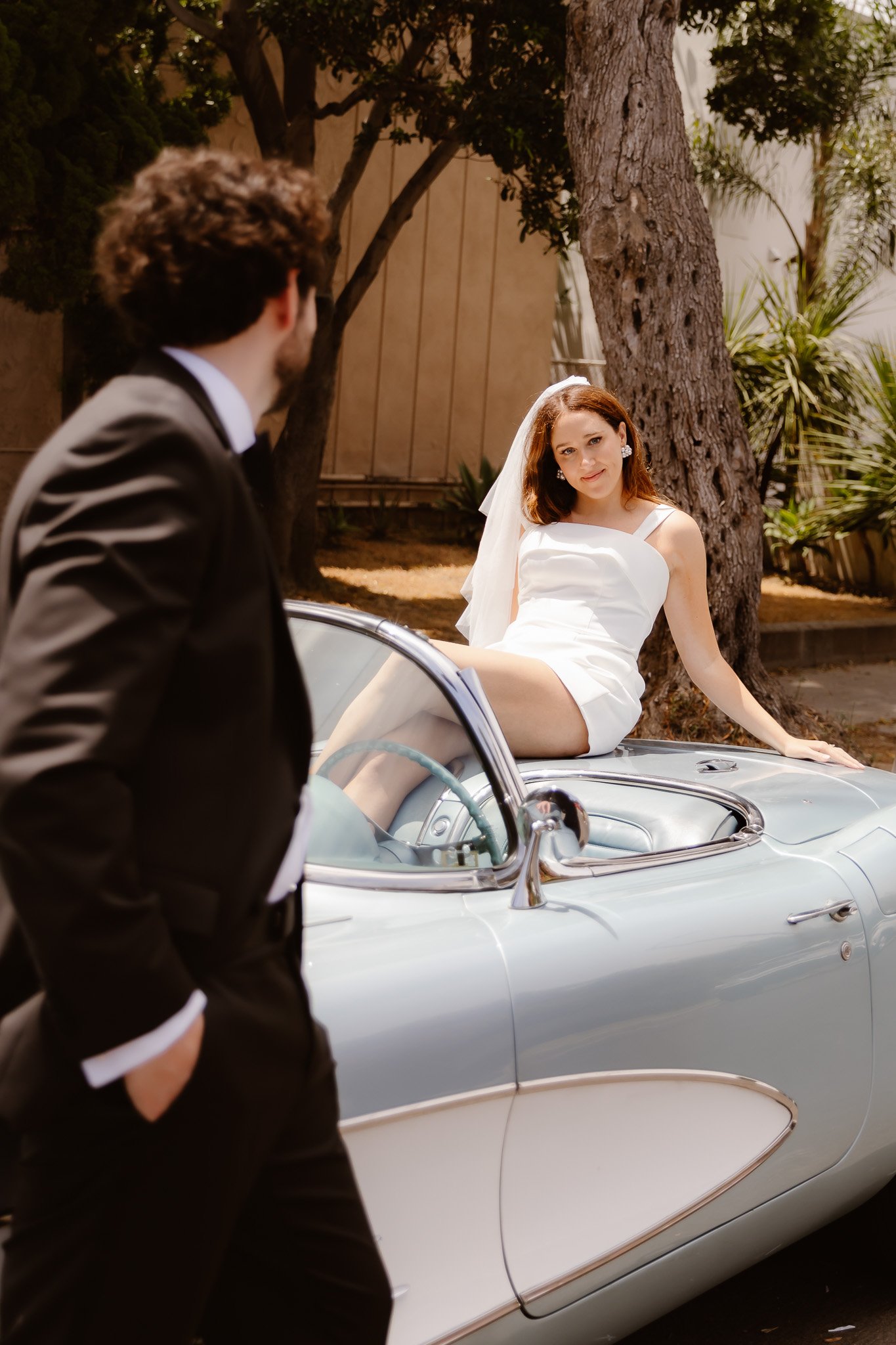 A woman in a white dress and veil sitting on a vintage silver convertible car, with a man in a black tuxedo standing nearby, outdoors with trees and a wooden fence.