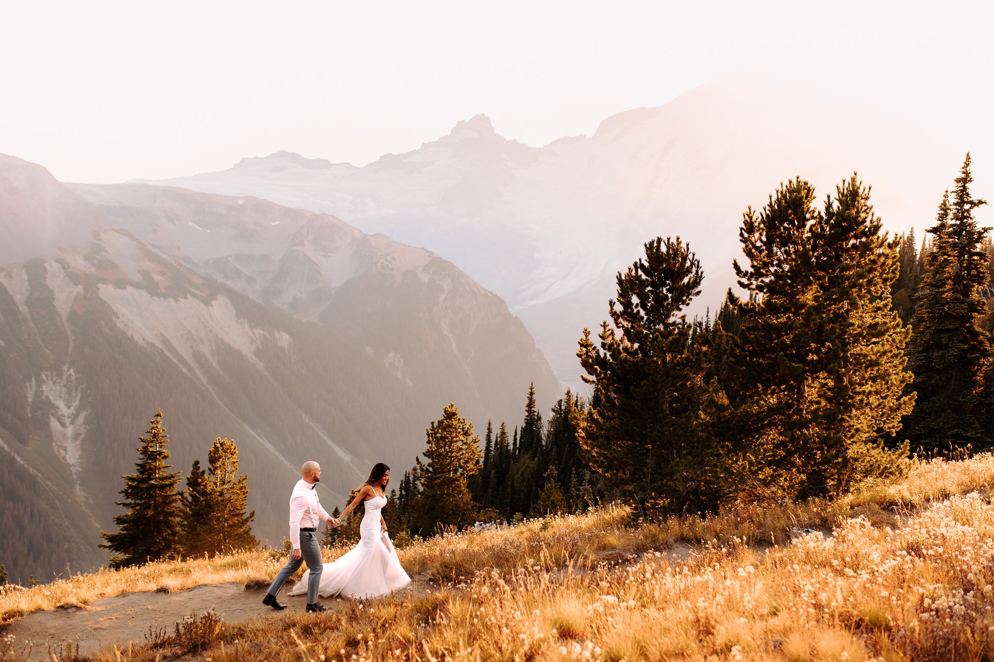 A couple holding hands walking on a trail through a mountain landscape at sunset, with trees and tall mountains in the background.