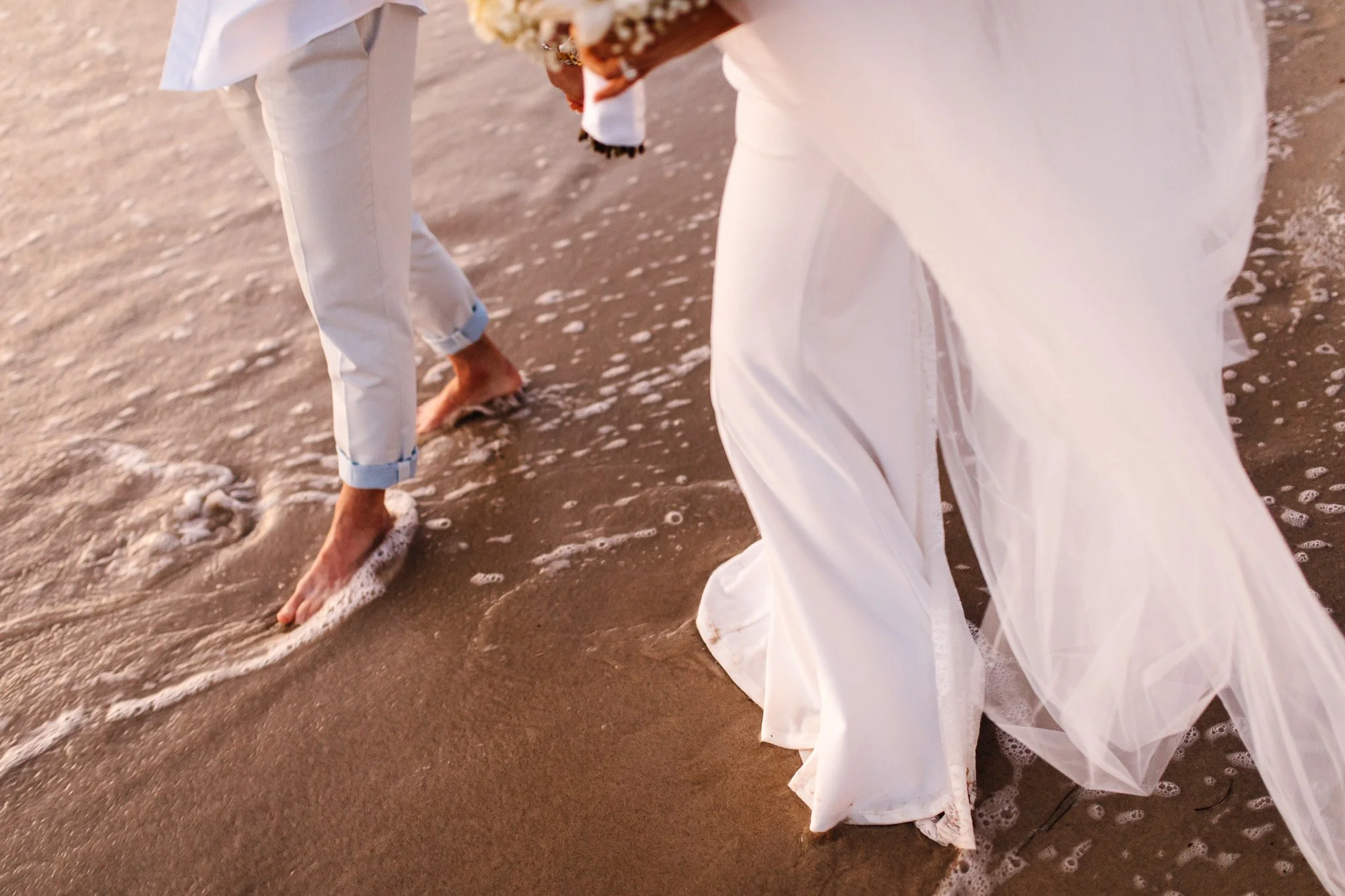 Two people standing barefoot on wet sand near the shoreline, wearing white clothing, with gentle waves washing over their feet, possibly at sunset or sunrise.