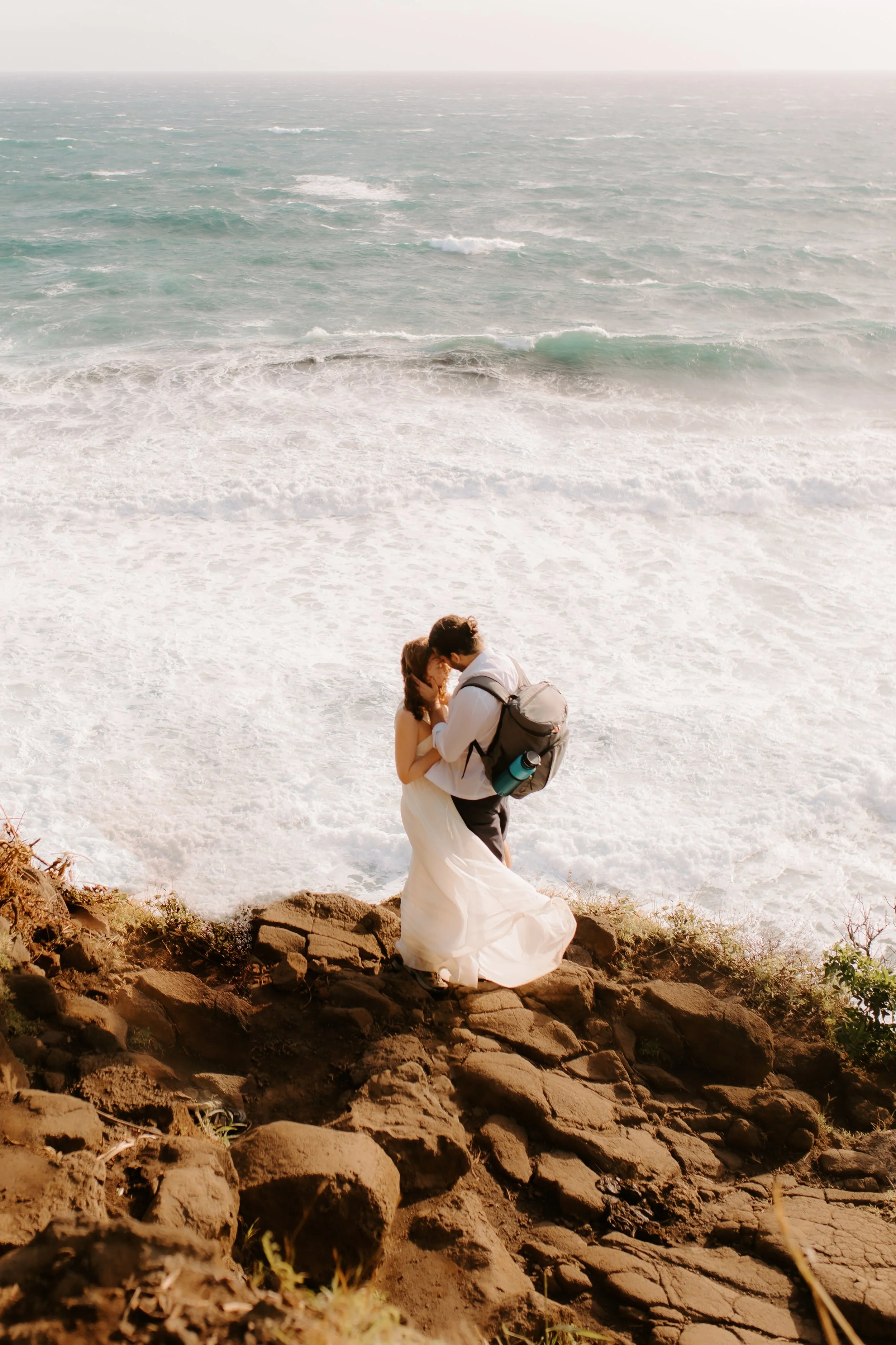 A couple dressed in wedding attire embracing and kissing on rocky terrain by the ocean, with waves crashing nearby.