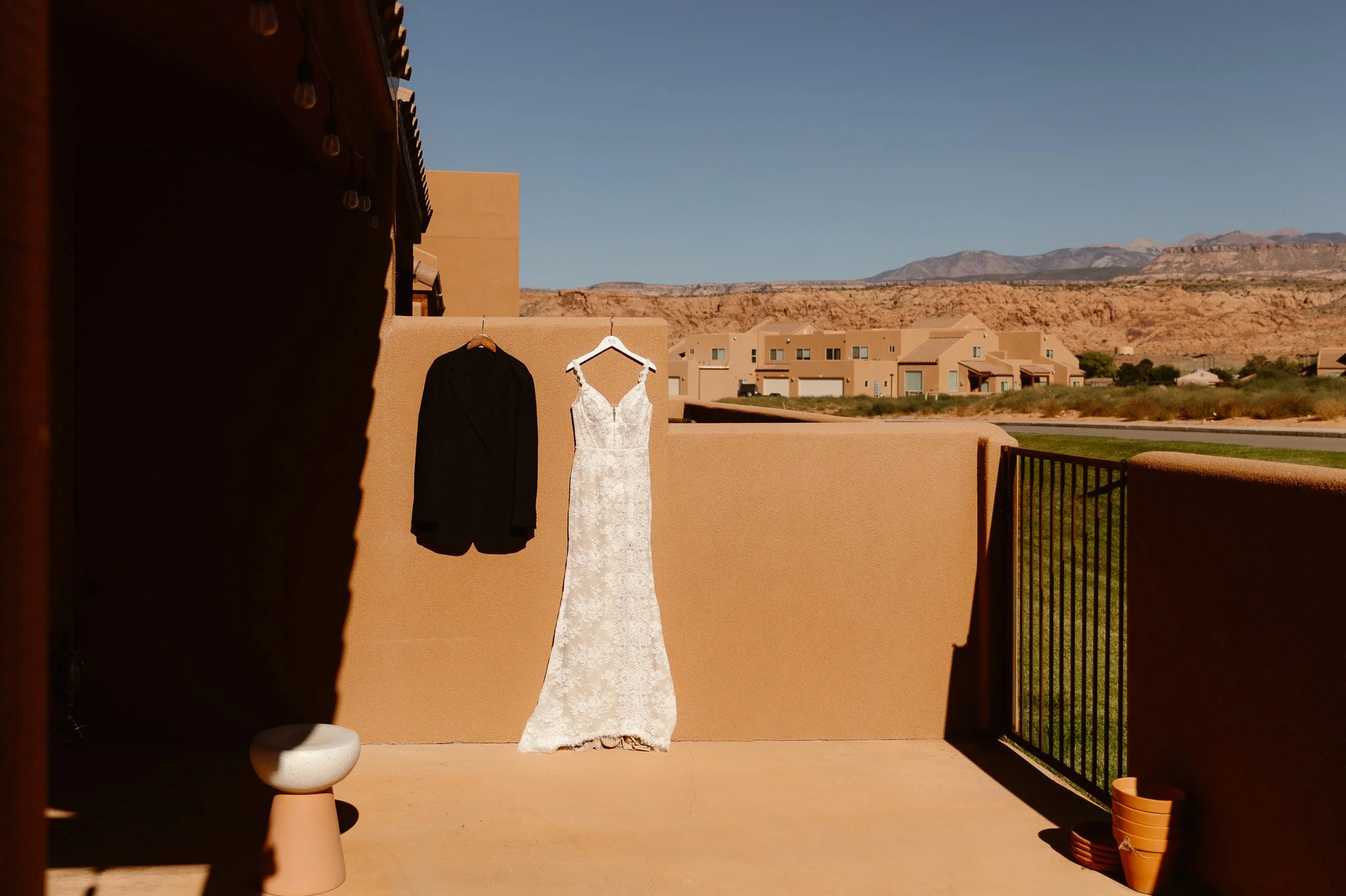 A wedding dress and a black suit hanging on a beige wall outdoors with desert mountains in the background, under a clear blue sky.