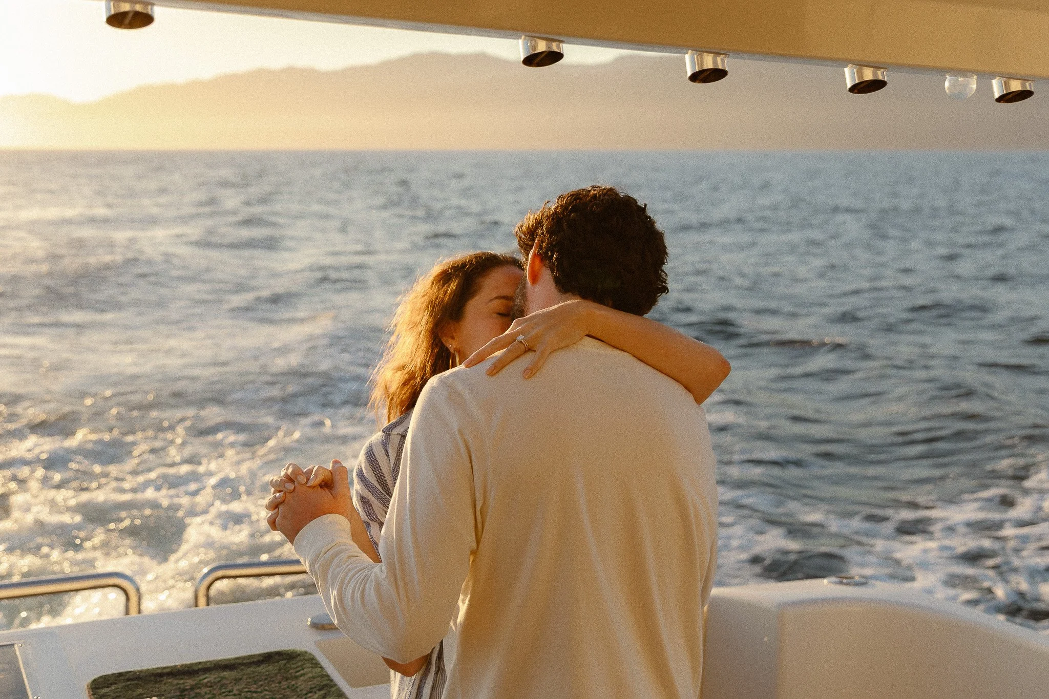 A couple dancing on a boat during sunset, with the ocean and mountains in the background.