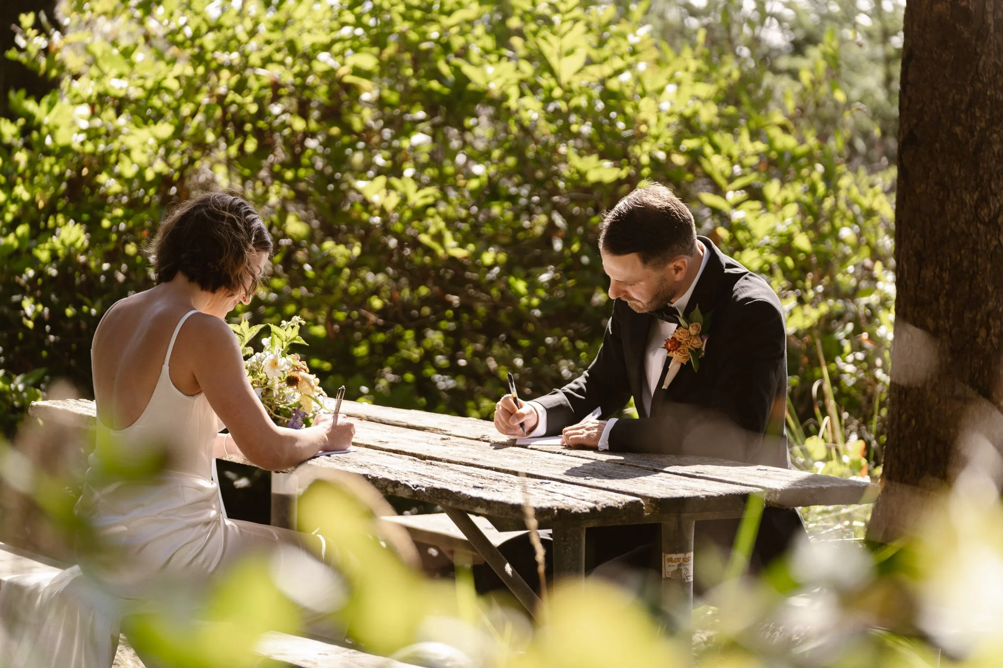 A bride and groom signing documents outdoors during their wedding ceremony, seated at a rustic wooden table surrounded by lush green foliage.