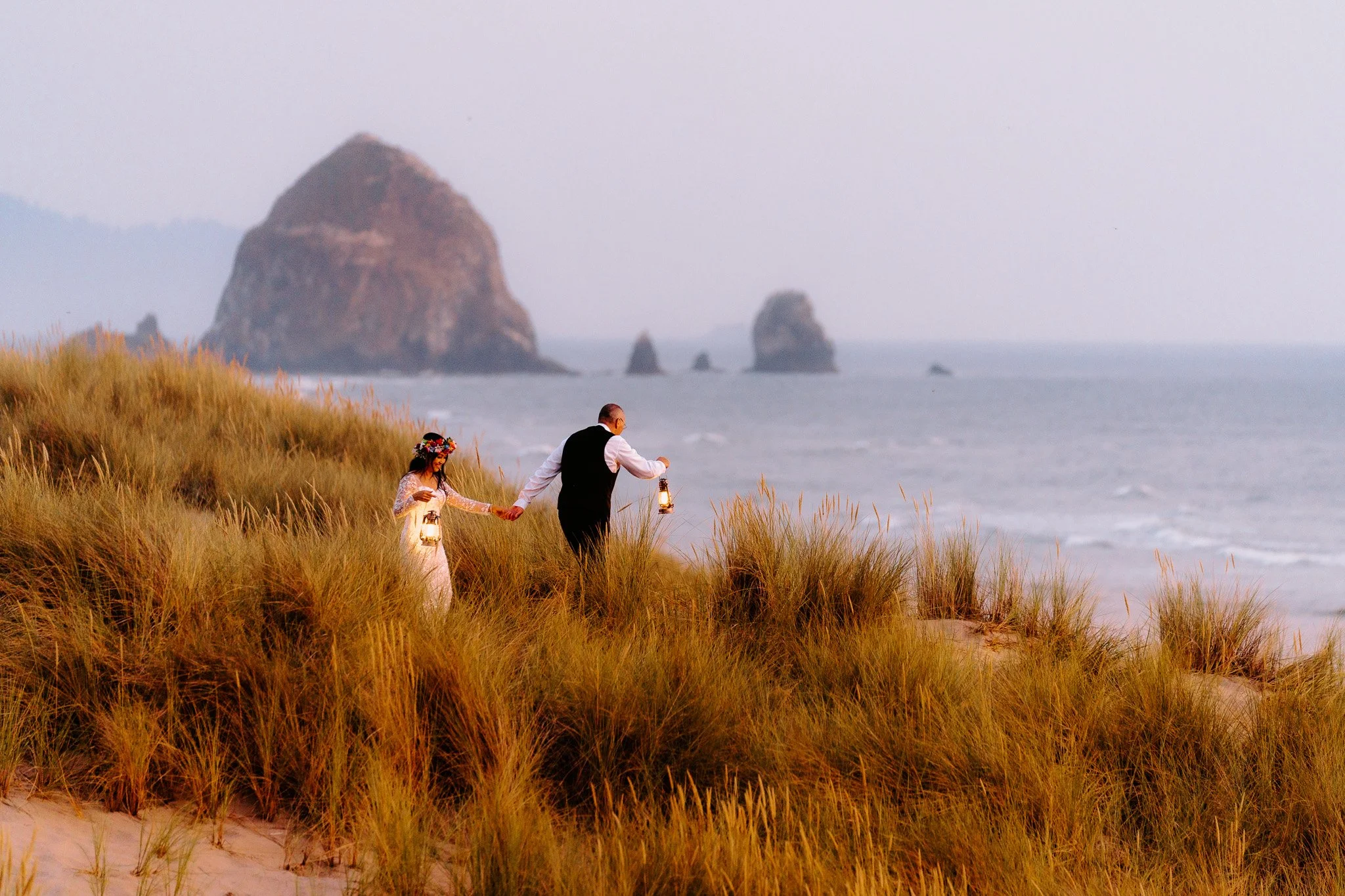 A couple walking on a grassy beach at sunset, holding lanterns, with large rock formations in the ocean in the background.