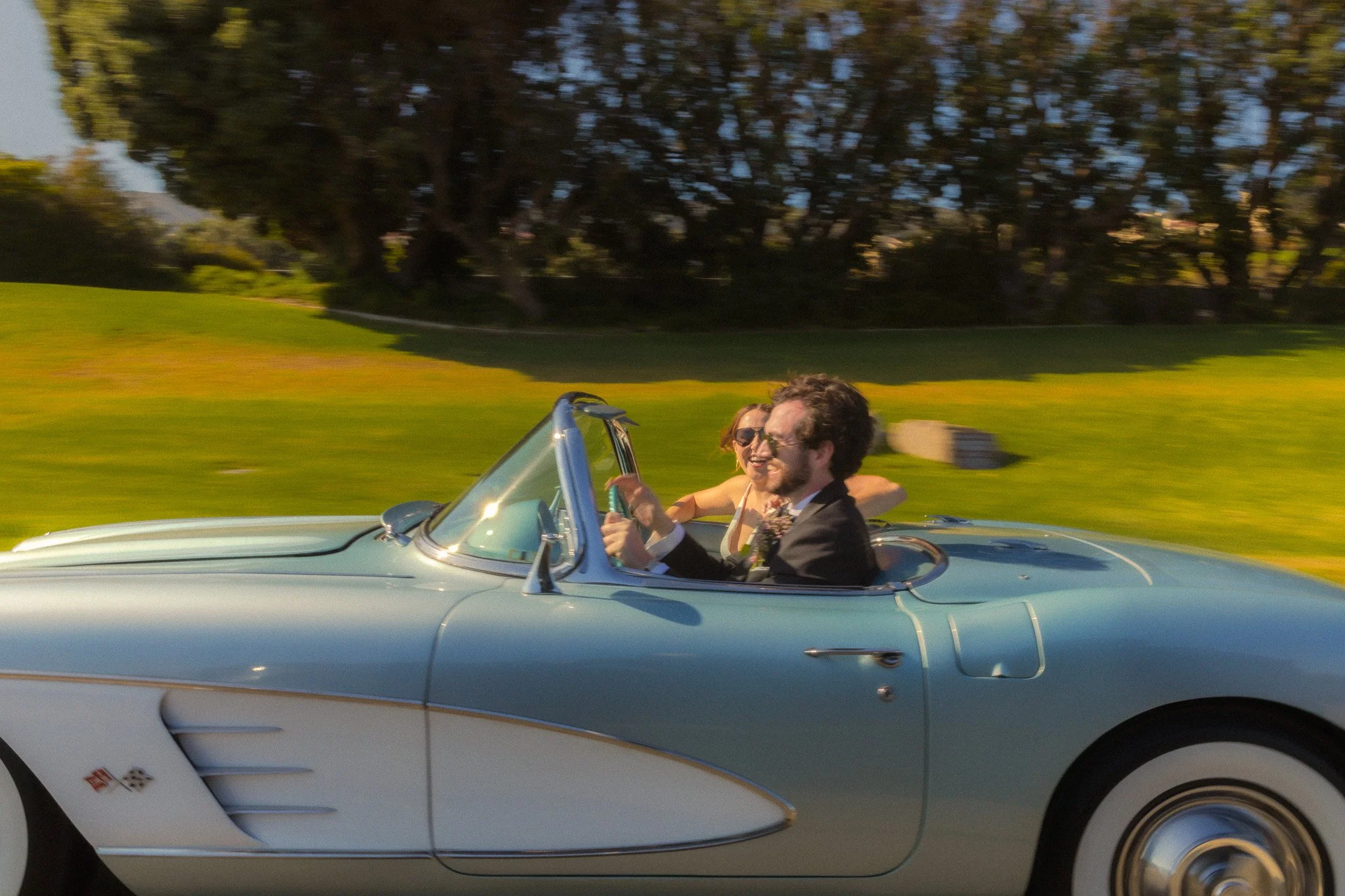 A classic silver convertible car with two people inside, driving down a road with green grass and trees in the background. The man in the driver's seat is wearing sunglasses and a suit, while the woman in the passenger seat is smiling and also wearin