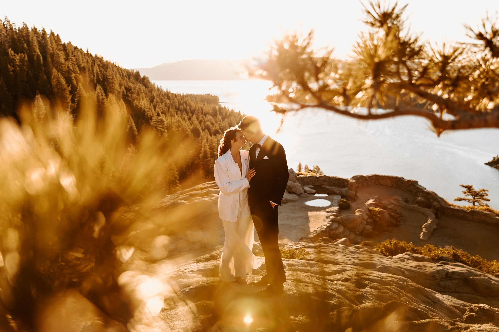 A couple in wedding attire sharing a kiss on a rocky beach at sunset, with a backdrop of a forested hillside and a lake.
