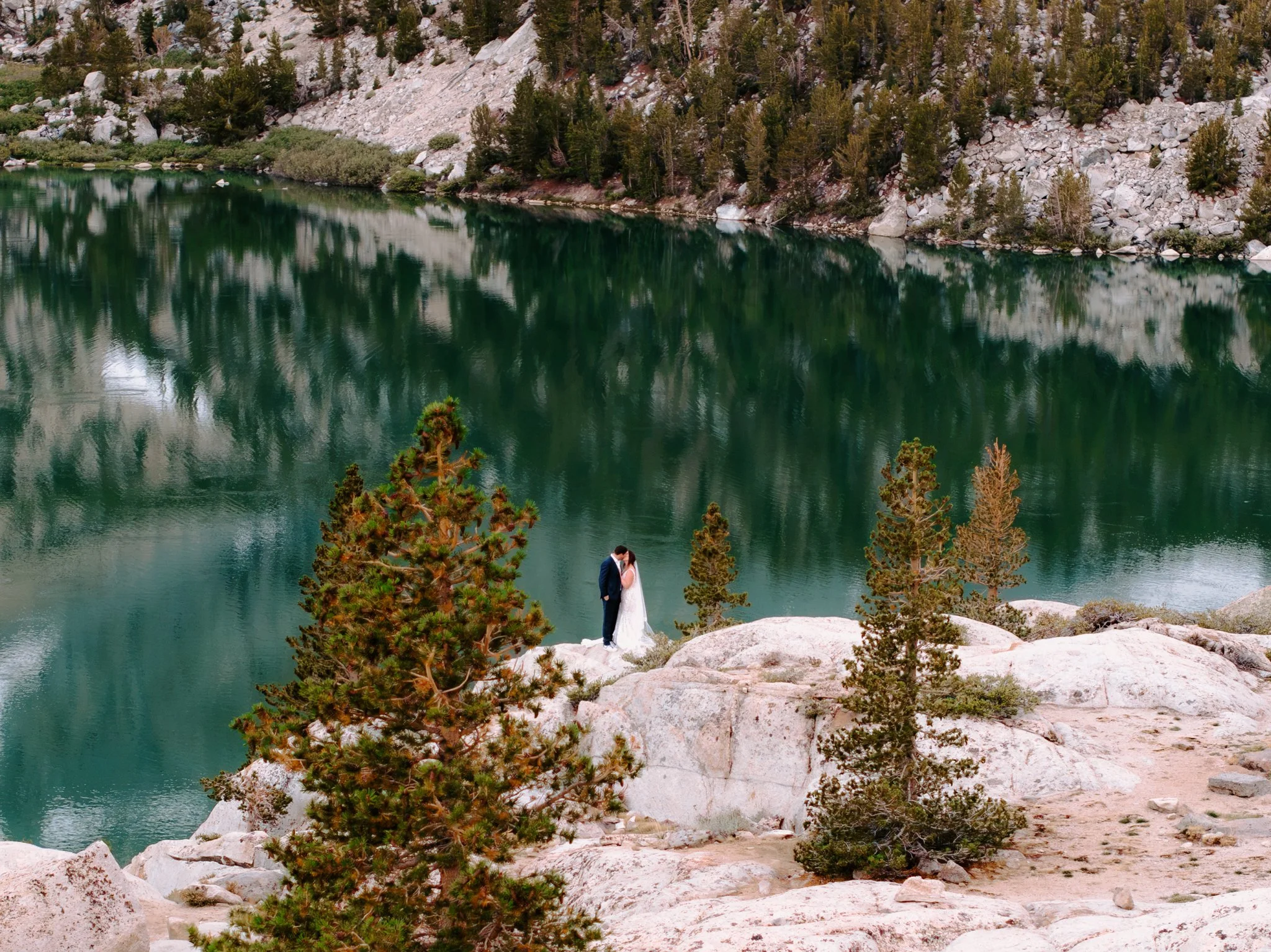 A married couple stands on a rocky ledge, surrounded by pine trees, with a green lake and rocky hillside in the background during daylight.