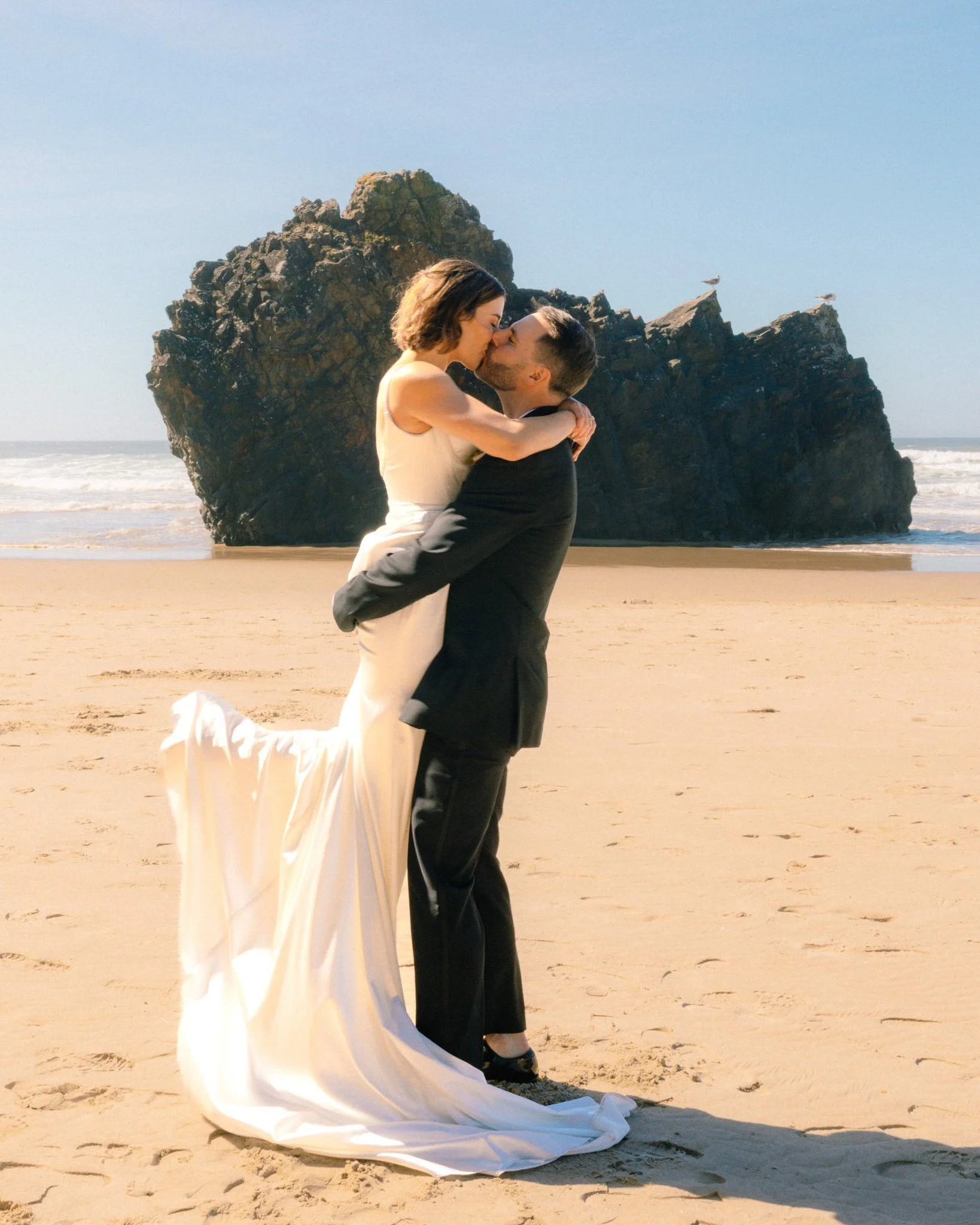 A couple dressed in wedding attire sharing a kiss on the beach with large rock formations and seagulls in the background.