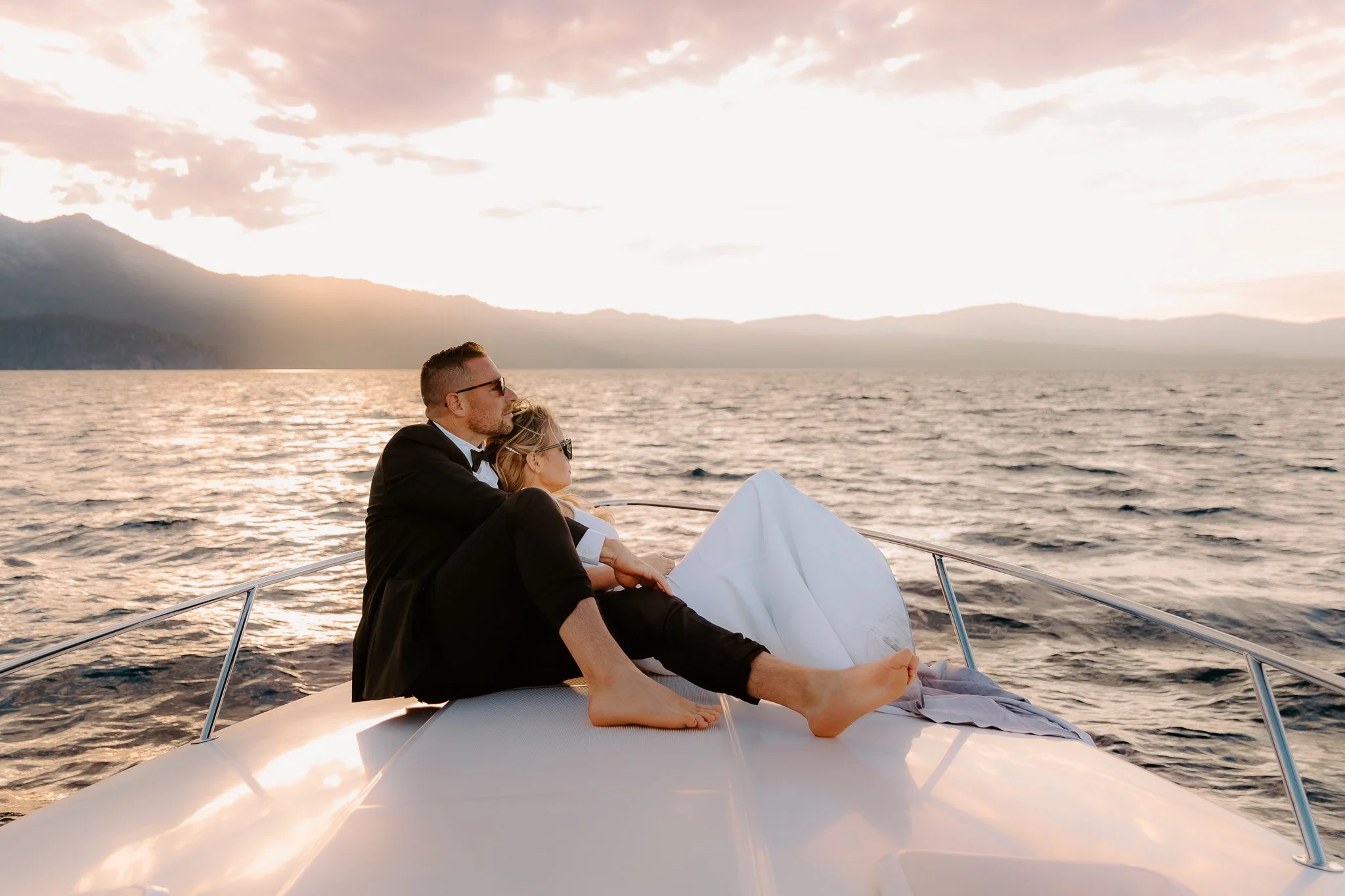 A couple dressed in formal attire, the man in a tuxedo and the woman in a wedding dress, sitting closely together on a boat at sunset, with a scenic view of mountains and water in the background.