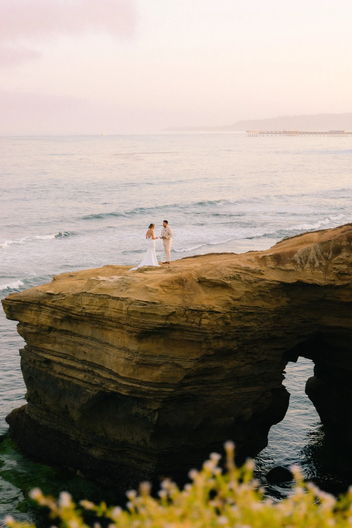 A couple in white wedding attire standing on large rock formations by the ocean, with waves at their feet and a distant pier and boat in the background.