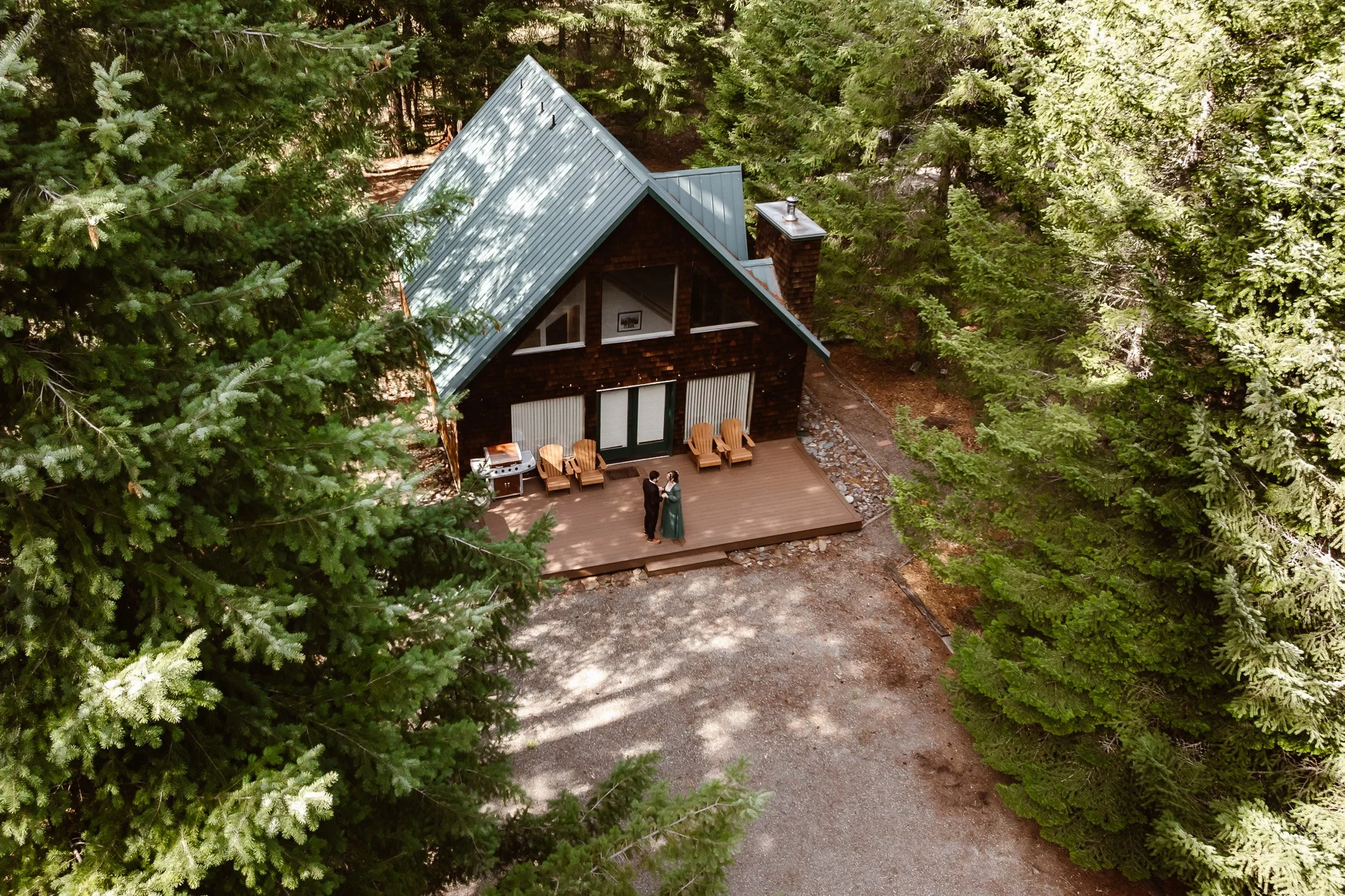 A house with a steep metal roof surrounded by tall green trees, with a wooden deck where two people are standing and talking.