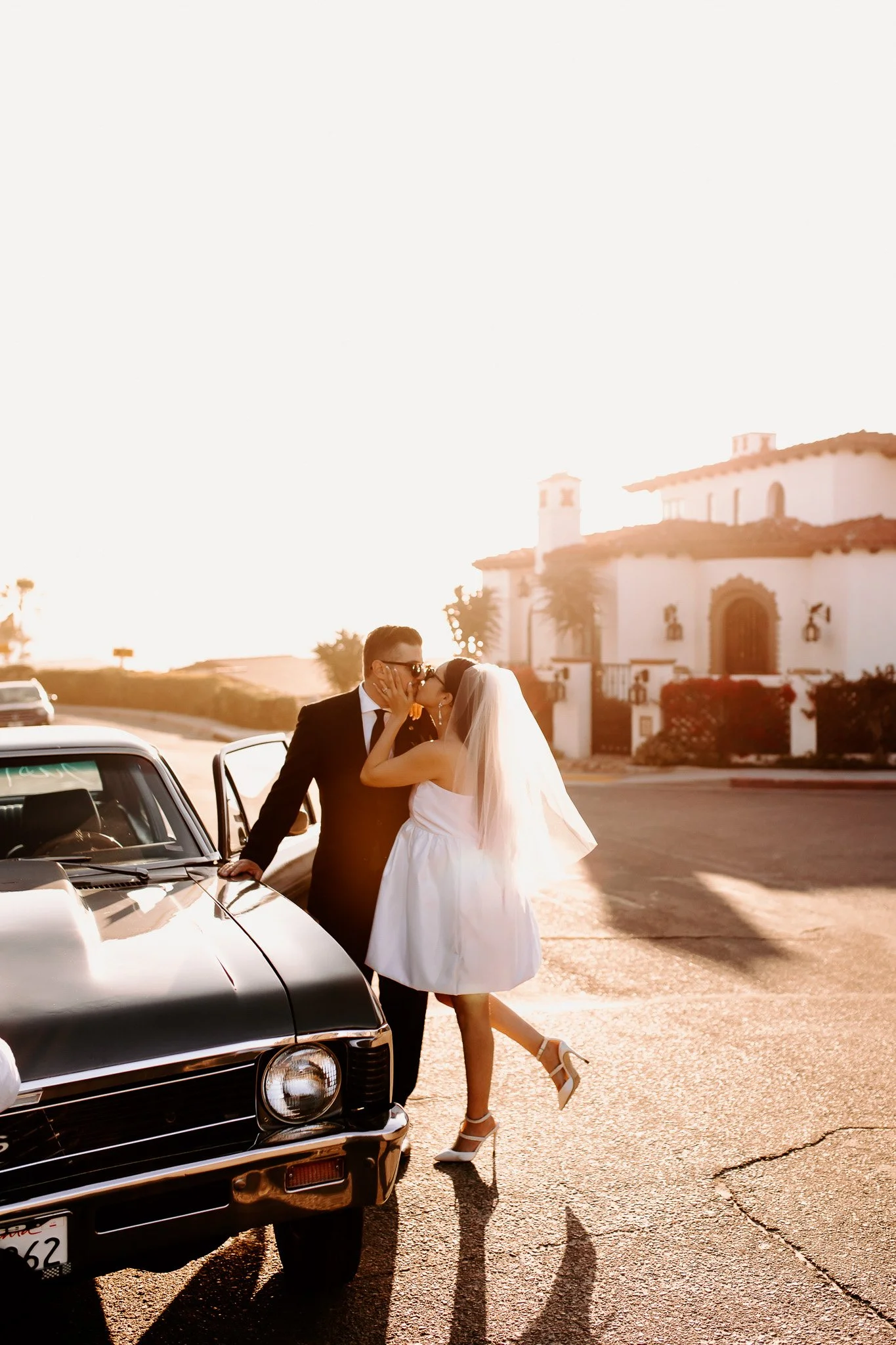 A bride and groom sharing a kiss outdoors at sunset, with a white wedding dress and black tuxedo, near a classic black car, in front of a large white building with arches and palm trees.