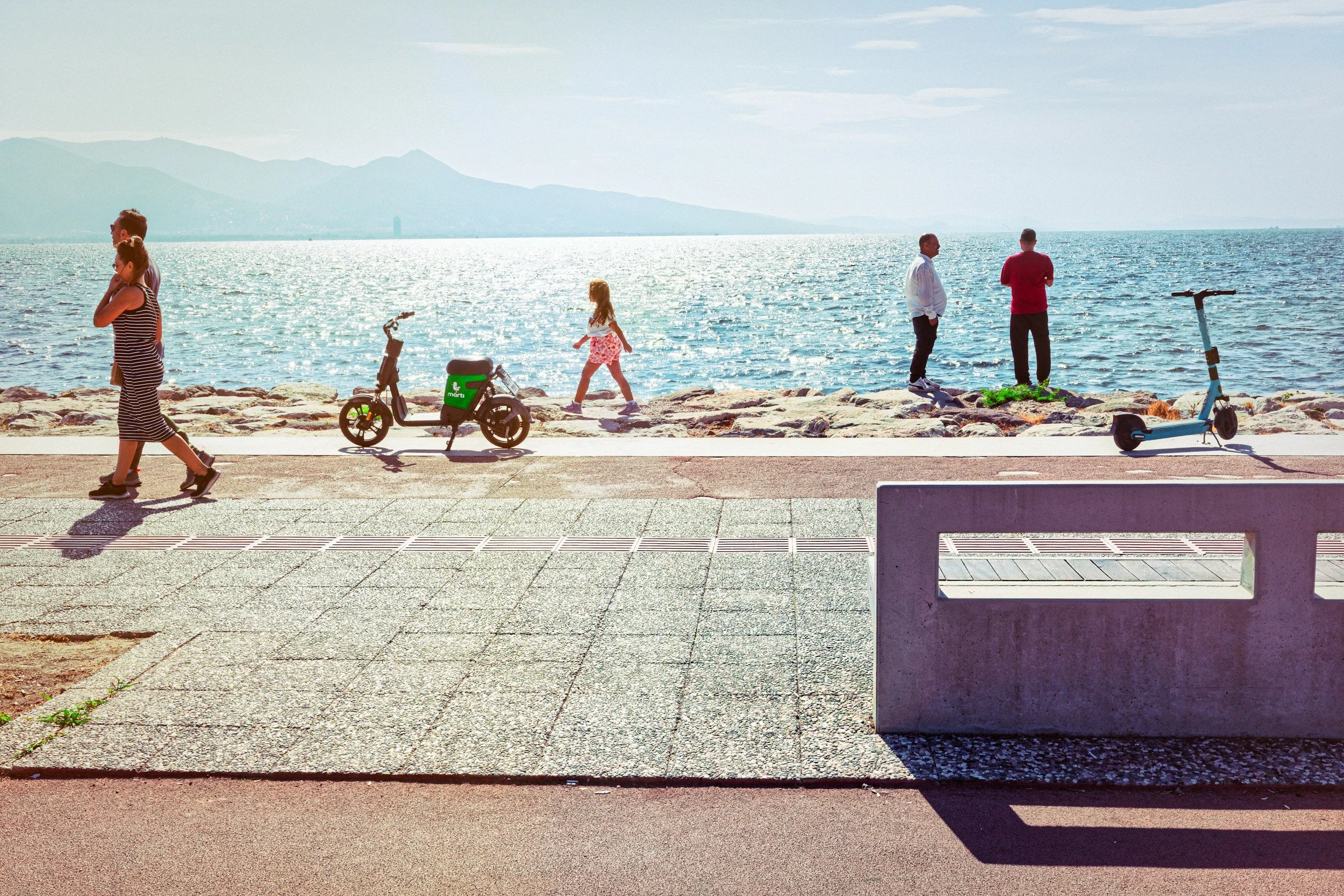 People walking and standing along a seaside promenade, with mountains and the ocean in the background and electric scooters parked nearby.
