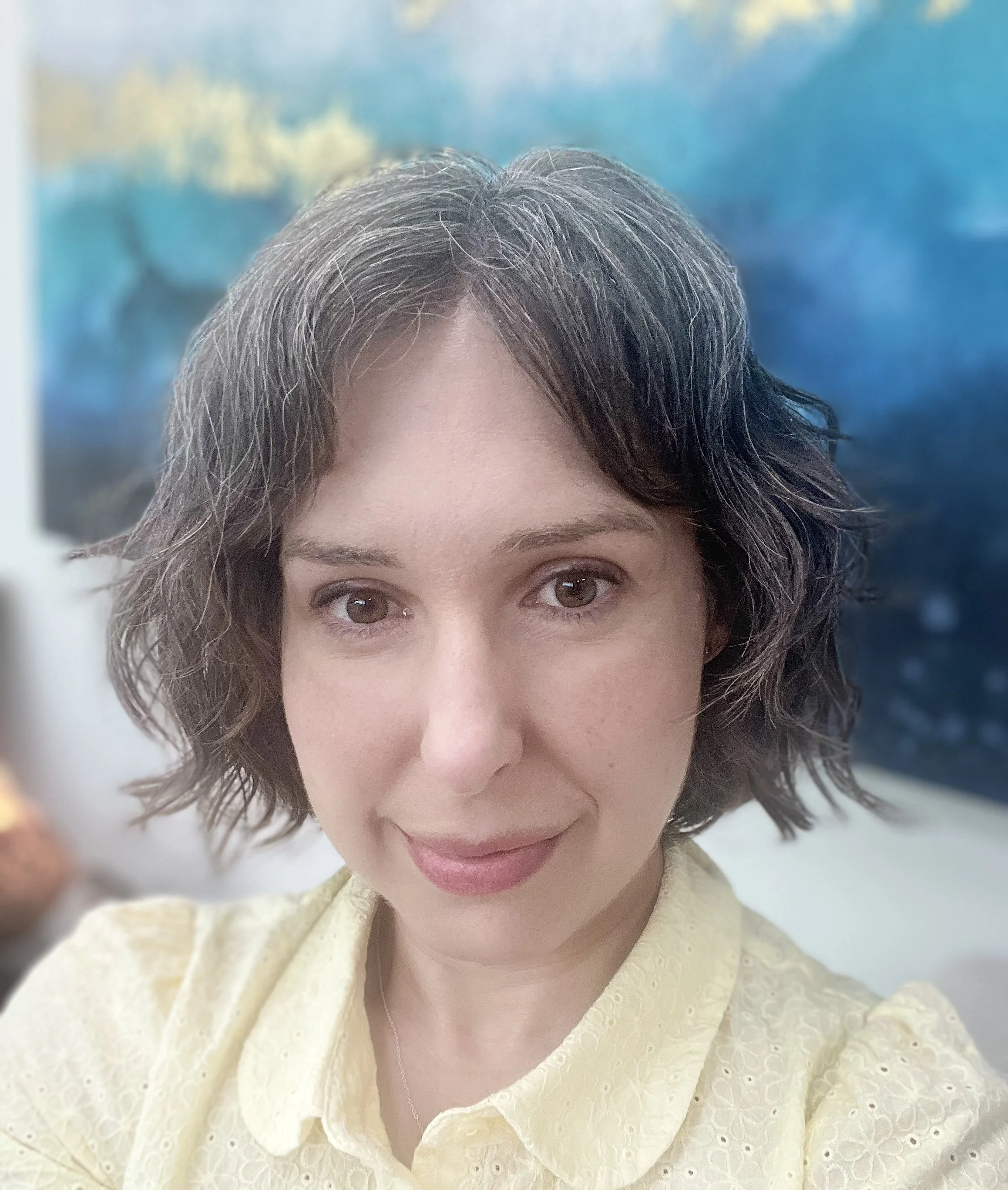 Close-up of a woman's face with short, wavy, grayish-brown hair, brown eyes, wearing a yellow textured dress, in front of a colorful blurred background.