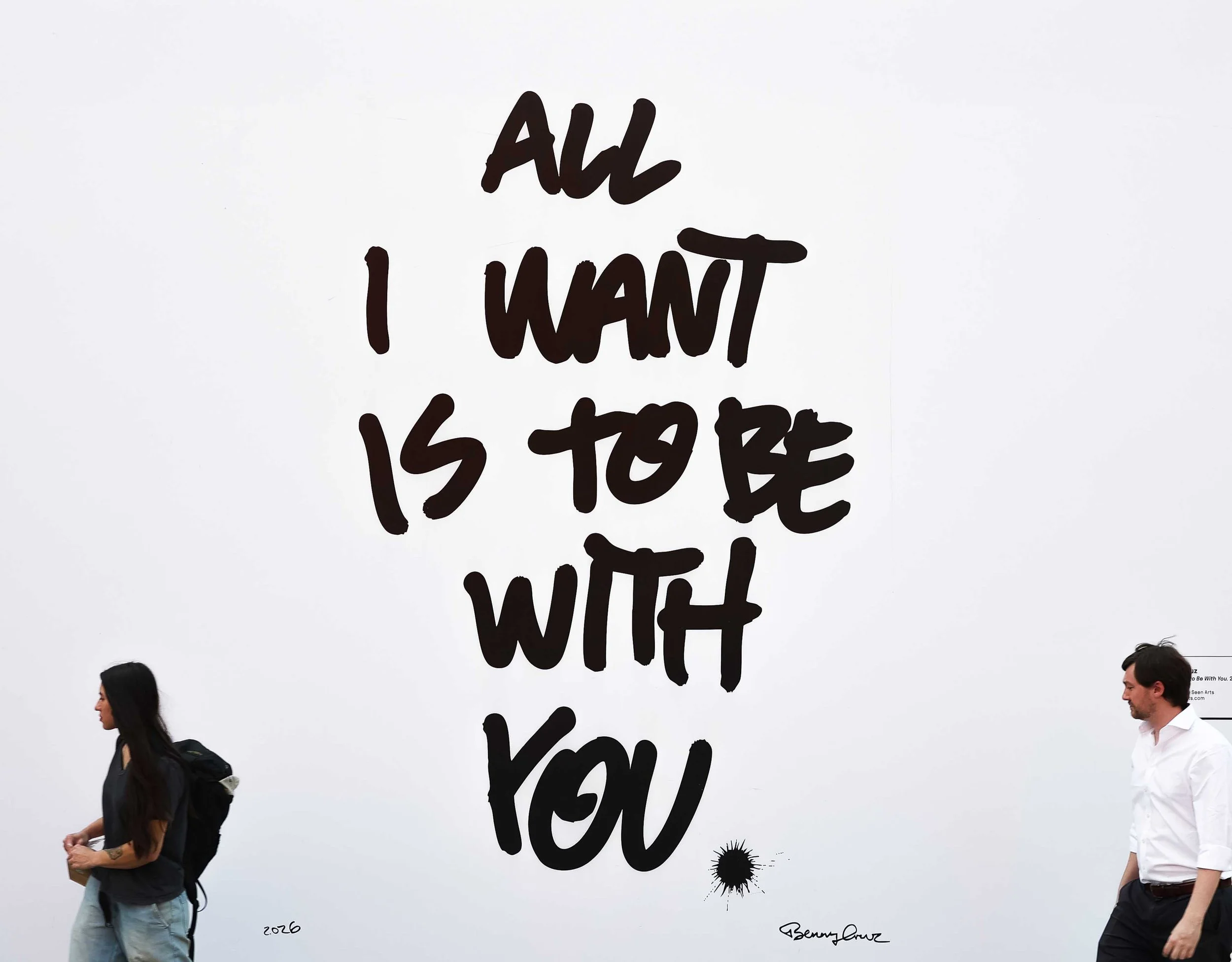 Pedestrians walking in front of Benny Cruz artwork that says "All I Want is to Be With You"