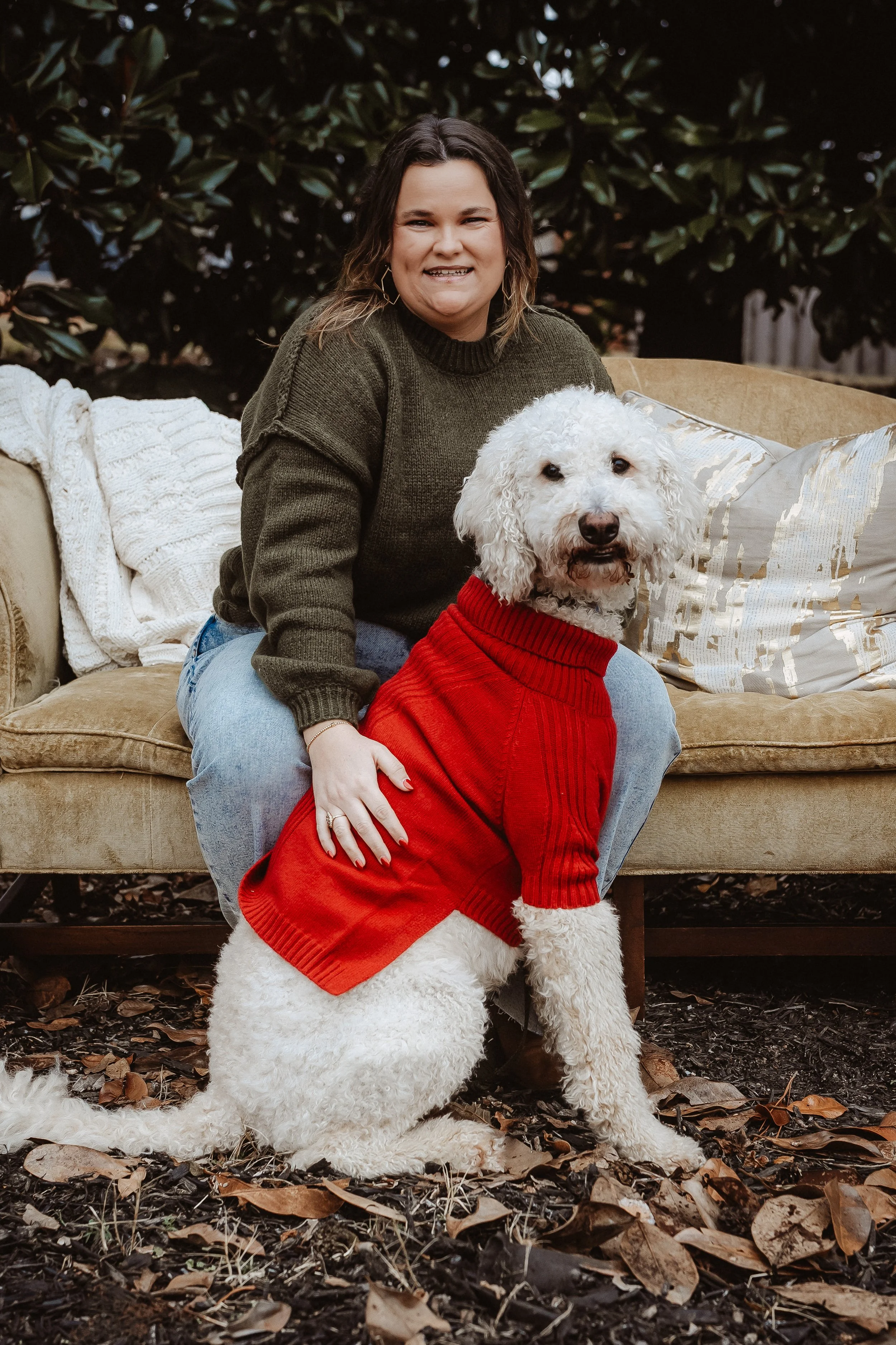 A woman wearing a green sweater and light blue jeans is sitting on a beige couch outdoors, smiling. A white dog with curly fur, wearing a red sweater, is standing in front of her, facing the camera, on a ground covered with fallen leaves.