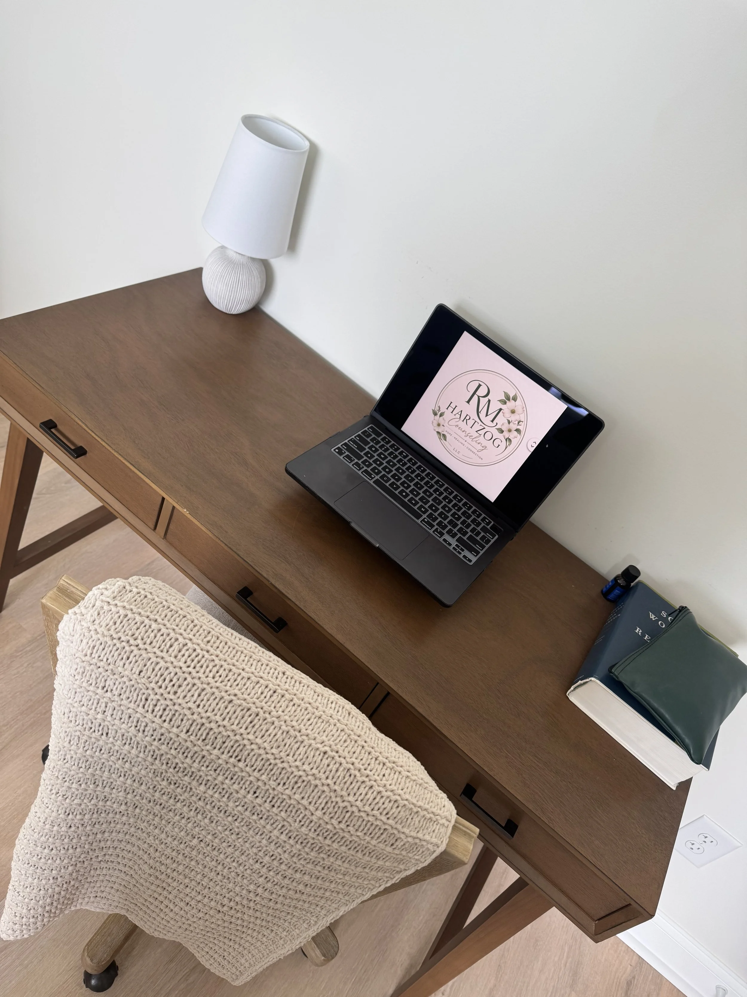 A wooden desk with a closed laptop displaying a logo for 'RM Hartzog Counseling' and a floral design, a blue notebook, a small blue bottle, a book, and a green wallet on the right side. A beige knitted office chair is in front of the desk.