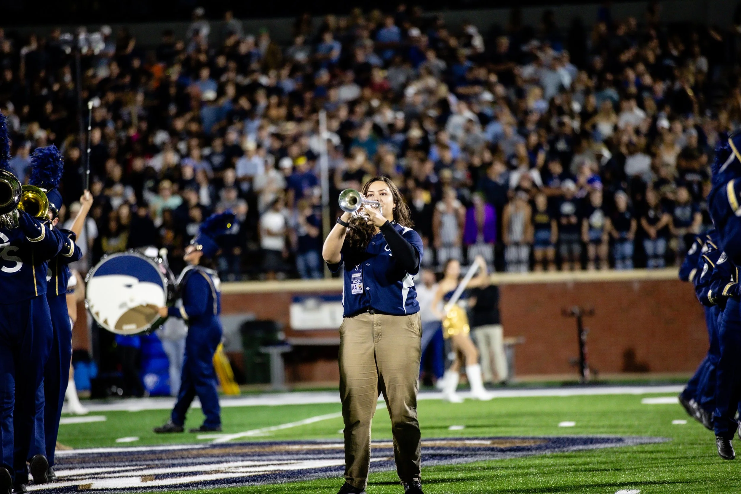 A woman playing the trumpet on a sports field during a night game with a large crowd in the stands. She is wearing khaki pants and a blue shirt, surrounded by band members in uniforms. Let's go Eagles!