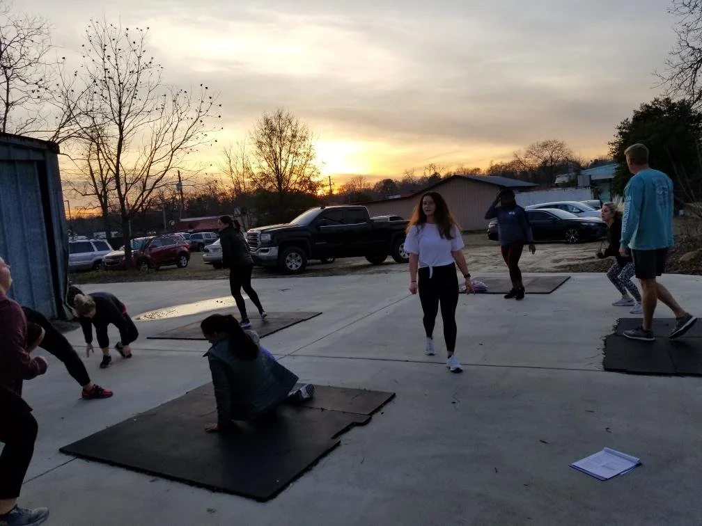 Group of people exercising outdoors on mats at sunset, with trees, parked cars, and a building in the background.