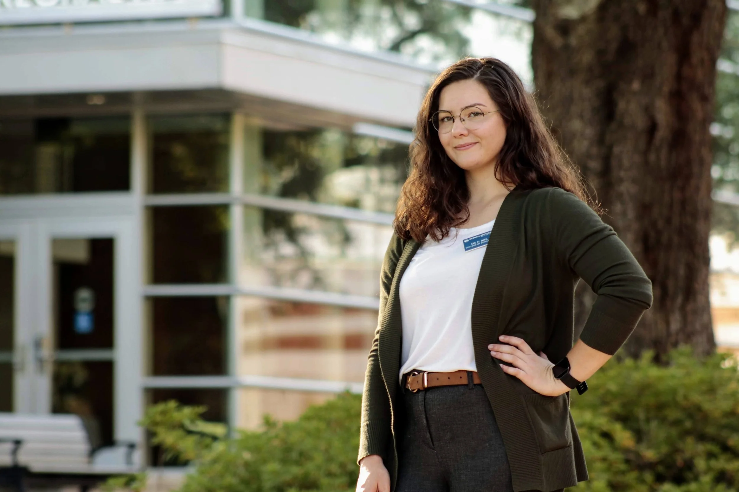 A woman with long curly brown hair and glasses standing outside next to a large tree, smiling with her hand on her hip, wearing a black blazer, white top, and a name tag.