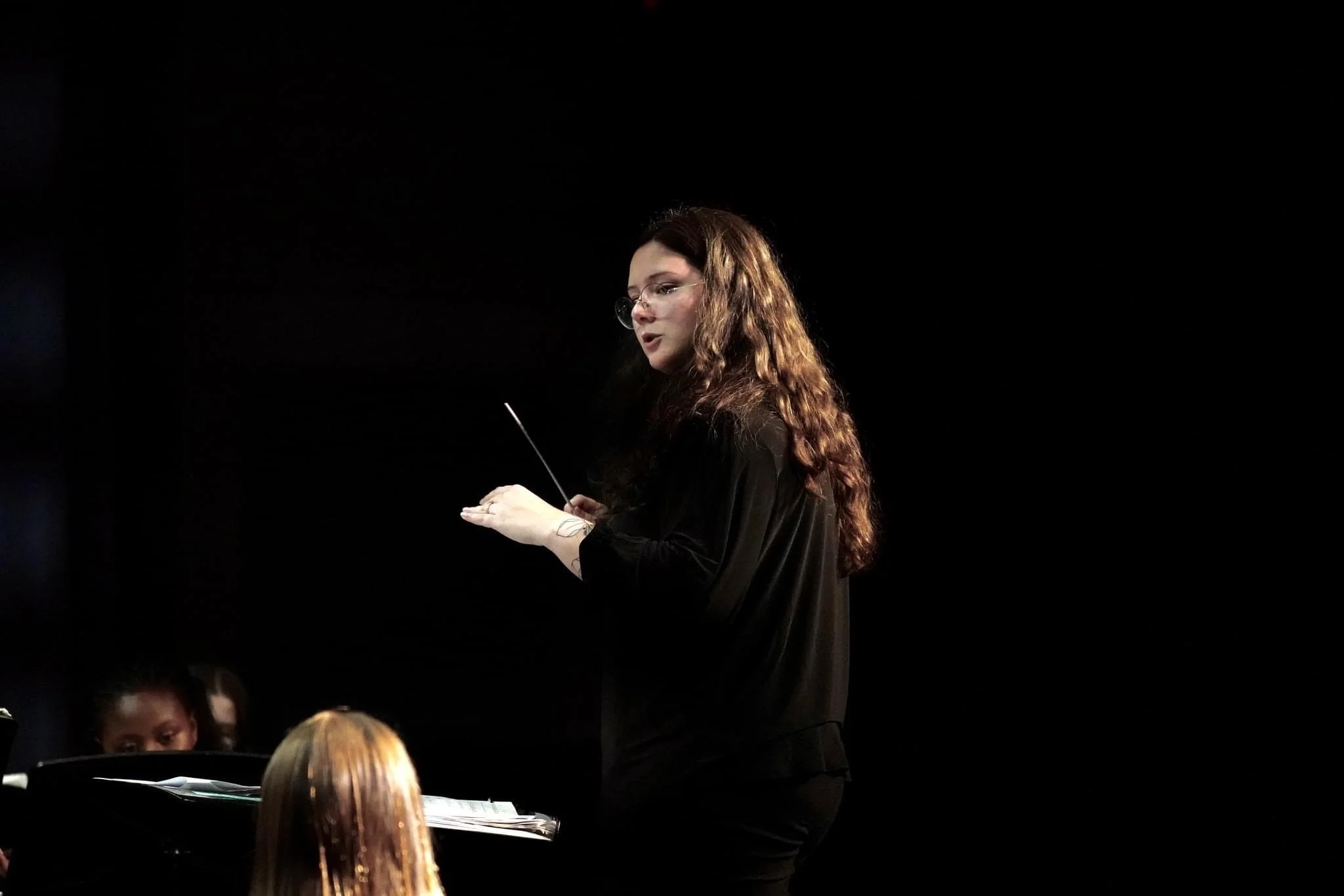 A woman with long curly brown hair and glasses conducts a wind ensemble, holding a baton, with a dark background and musicians visible in the foreground.