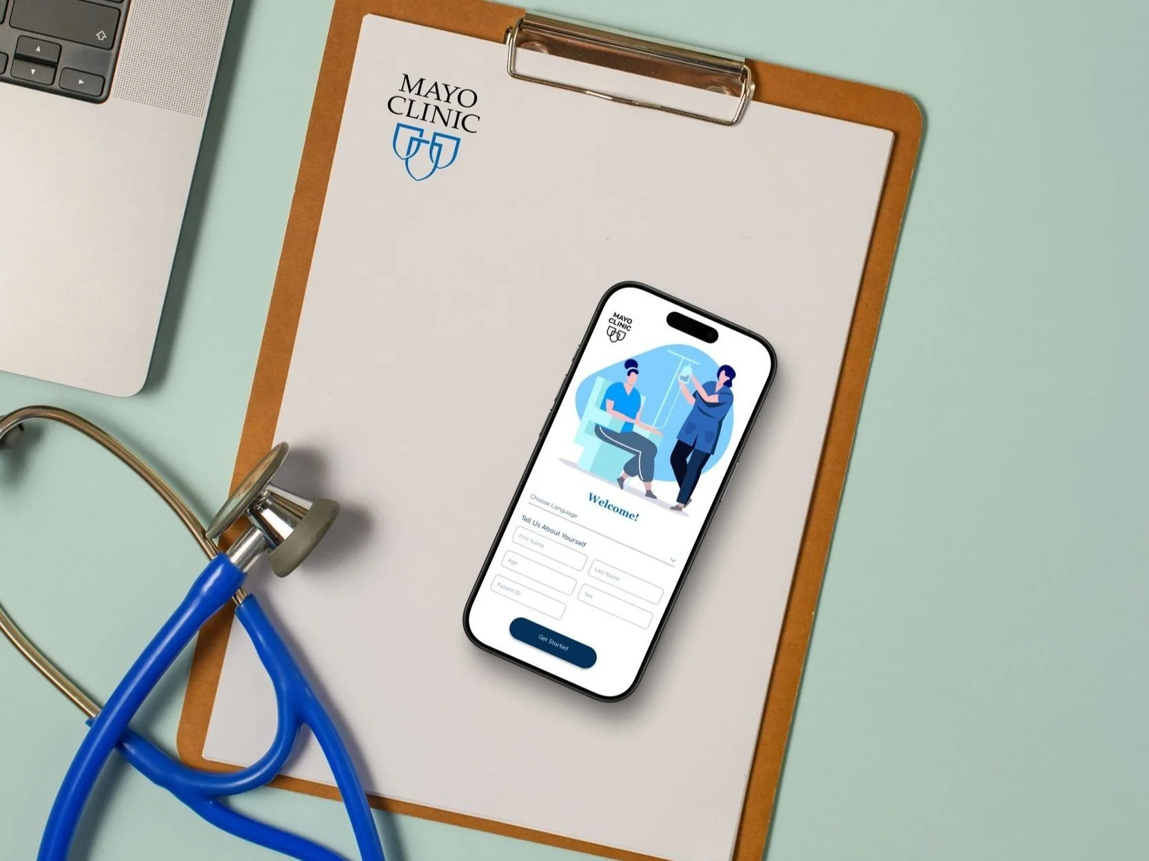 A medical clipboard with the Mayo Clinic logo, an Apple iPhone showing a healthcare app, a stethoscope, and a partial view of a laptop on a light blue surface.