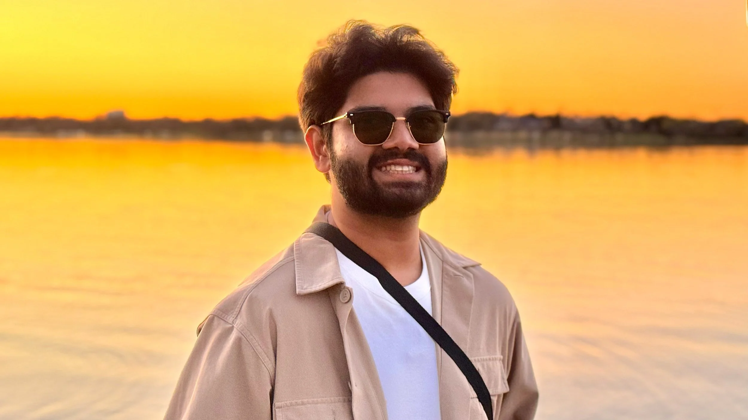 Man with sunglasses smiling by a lake at sunset.