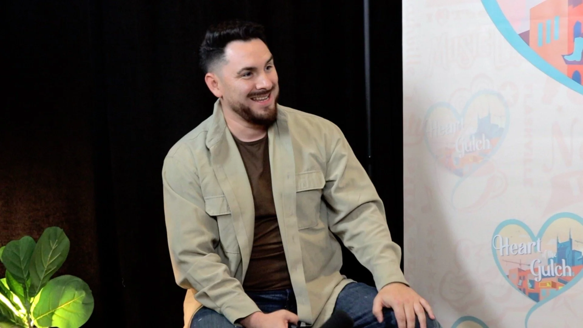 Tony Mendoza, host of Heart of the Gulch with dark hair, a beard, and light skin, smiling and sitting in front of a backdrop with the words 'Heart of the Gulch' and a heart-shaped logo, next to a green plant with large leaves.