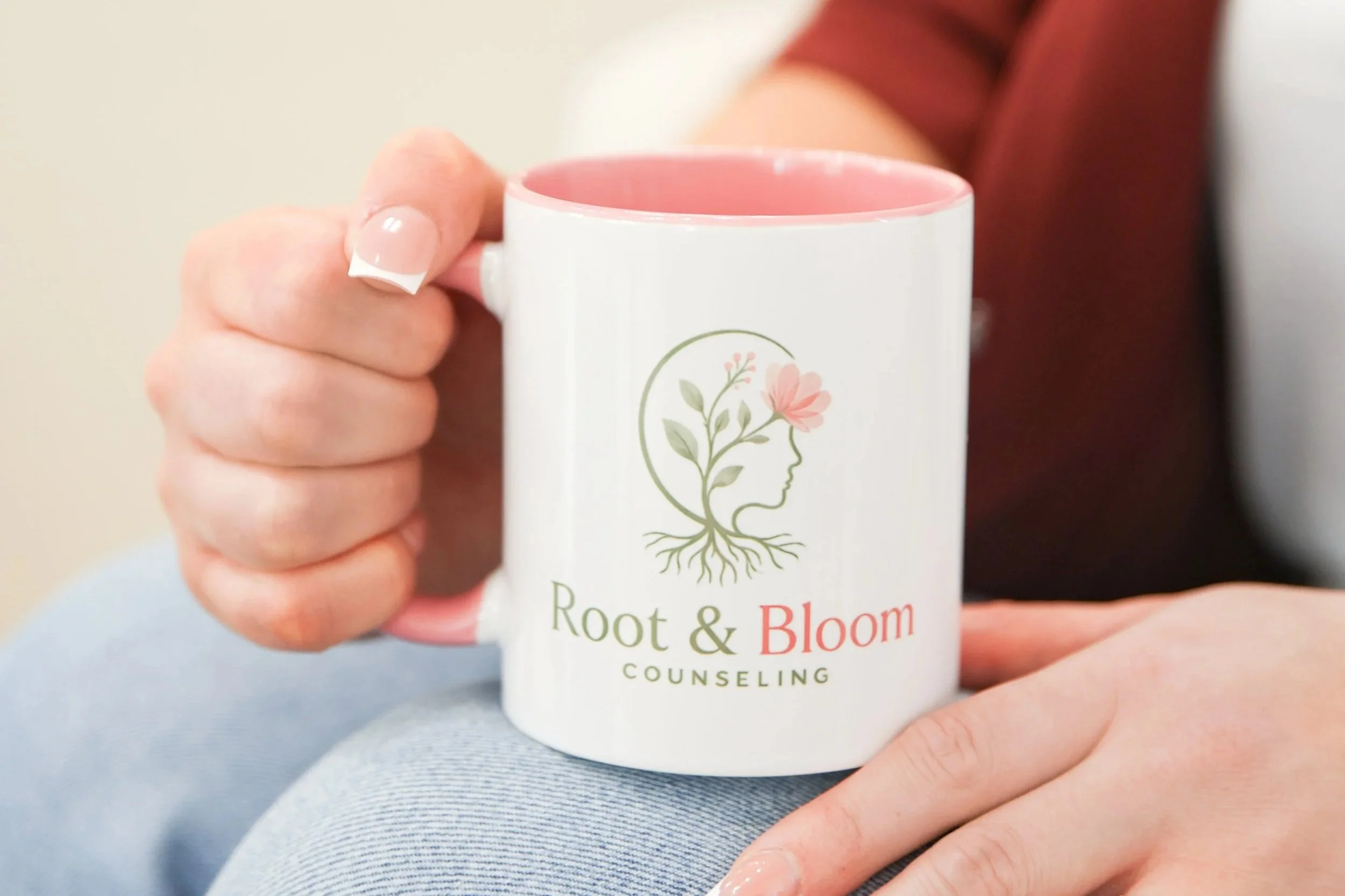 Close-up of a person holding a white ceramic mug with a pink interior. The mug features a logo with a girl's profile, floral elements, and the words 'Root & Bloom Counseling.' The person's hand has neatly trimmed nails with a light pink polish.