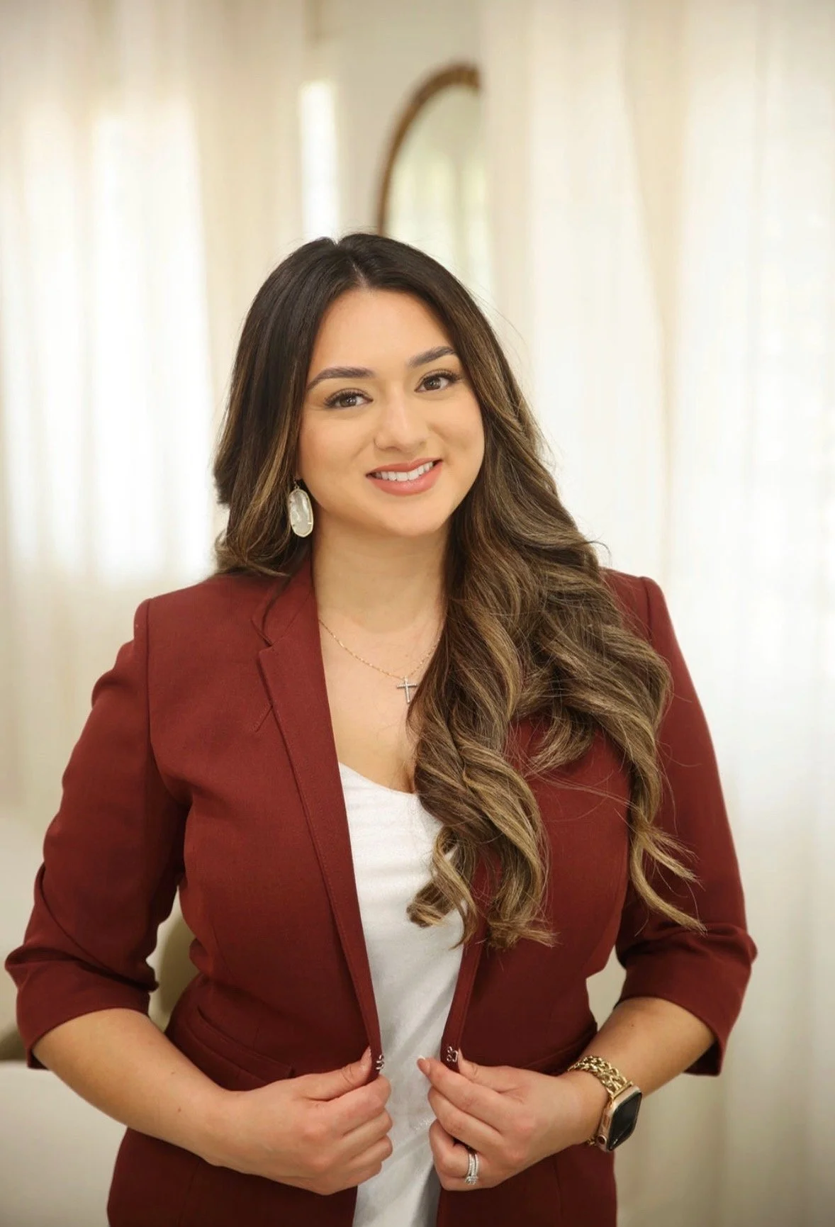Woman with long wavy hair in a maroon blazer, white top, wearing jewelry including earrings, rings, a necklace with a cross, and a watch, smiling in front of cream-colored curtains.