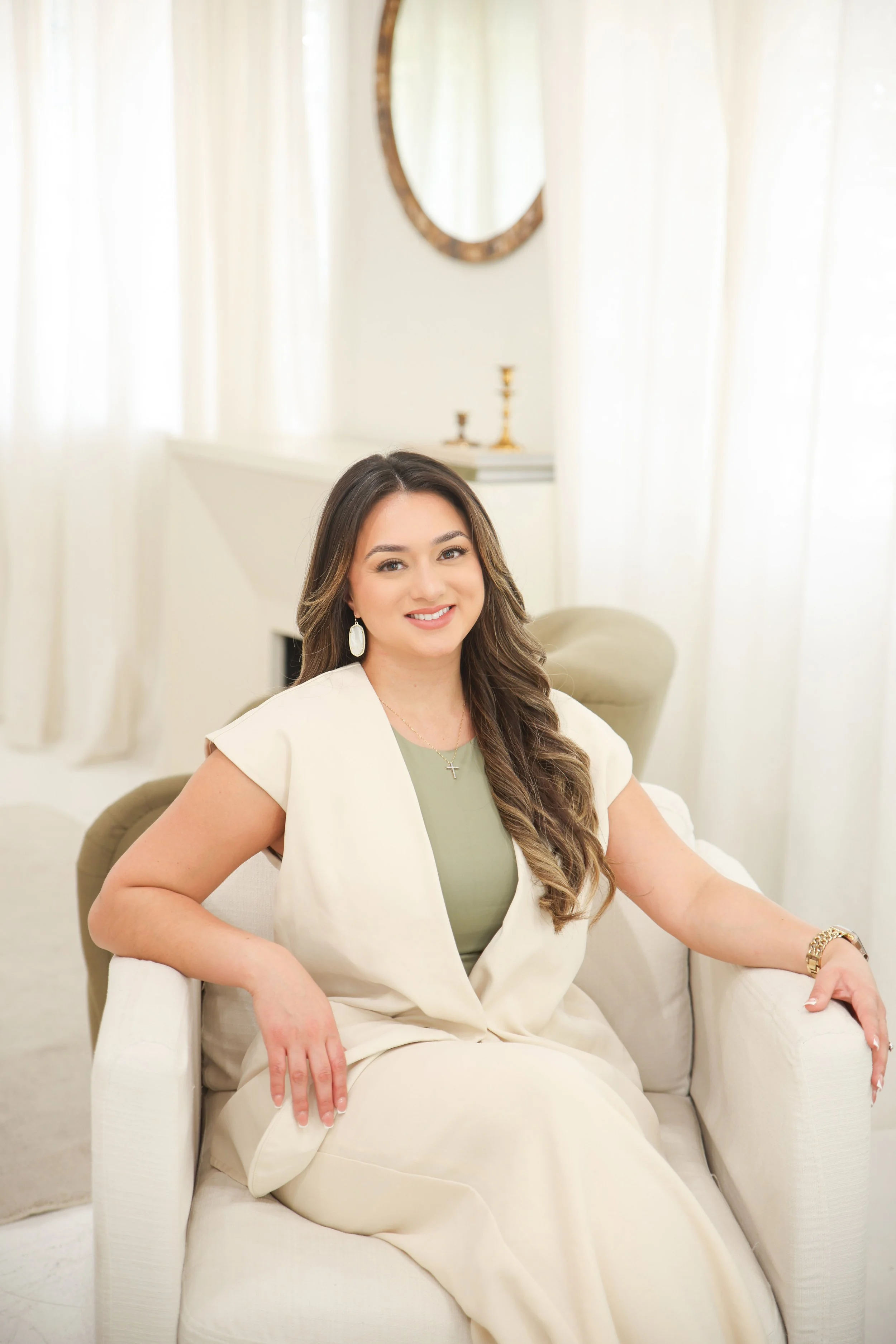 A woman with long brown hair, wearing a light green top and a cream-colored blazer, sitting on a cream-colored armchair in a bright, elegant room with white curtains, a mirror, and gold candlesticks in the background.