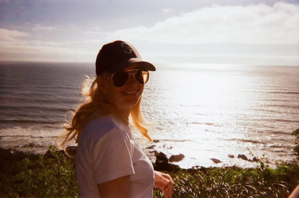 blonde girl in sunglasses and a Mariners hat smiling beside the ocean at sunset