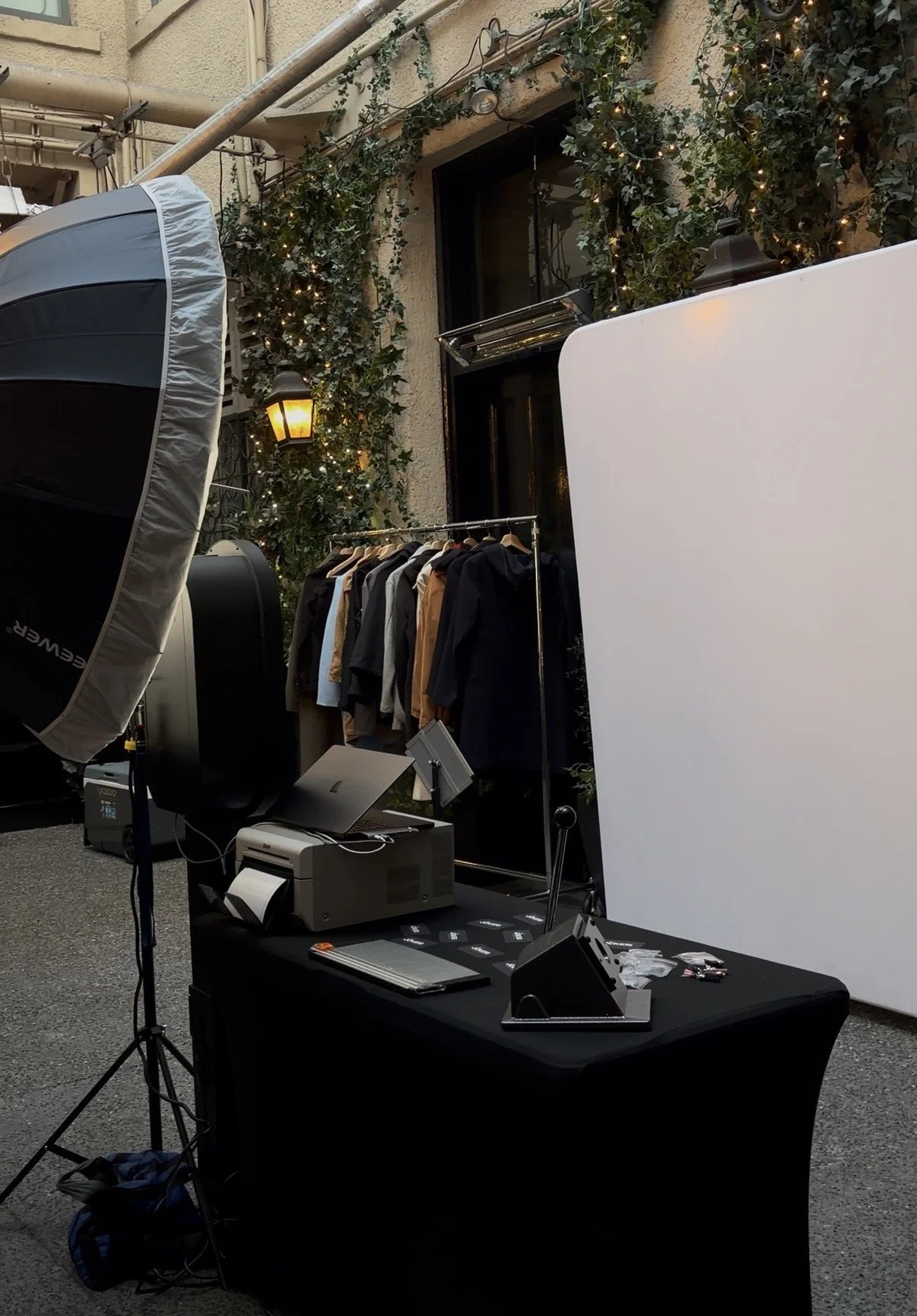 An outdoor photo of a clothing display at a photoshoot setup, with a rack of coats and jackets, a large white reflector, studio lights, and a table with photography equipment, set against a building decorated with string lights and ivy.