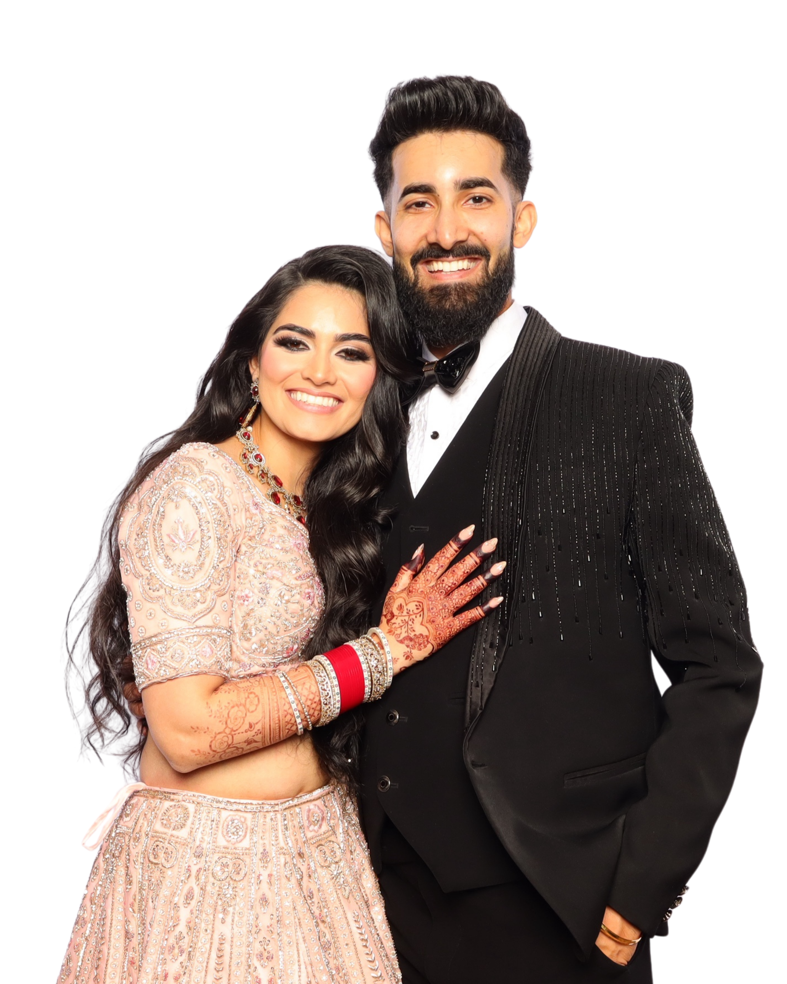 A smiling couple. The woman wears a traditional peach-colored Indian dress with embroidery, jewelry, and henna on her hands. The man wears a black tuxedo with a bow tie. They are standing close together against a white background.
