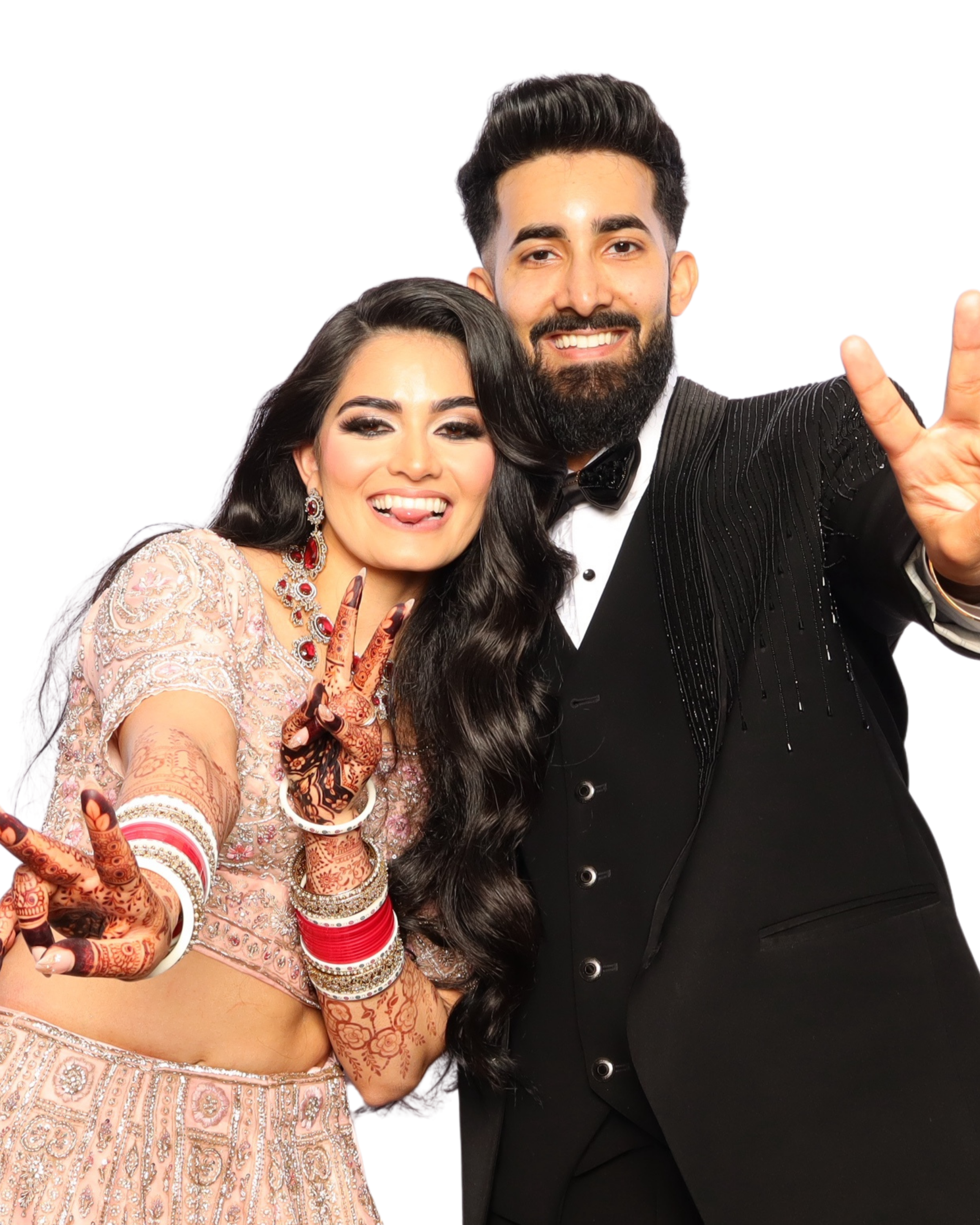 A man and woman taking a selfie, smiling and making playful gestures. The woman wears traditional Indian attire with henna on her hands, while the man wears a tuxedo, both against a plain white background.