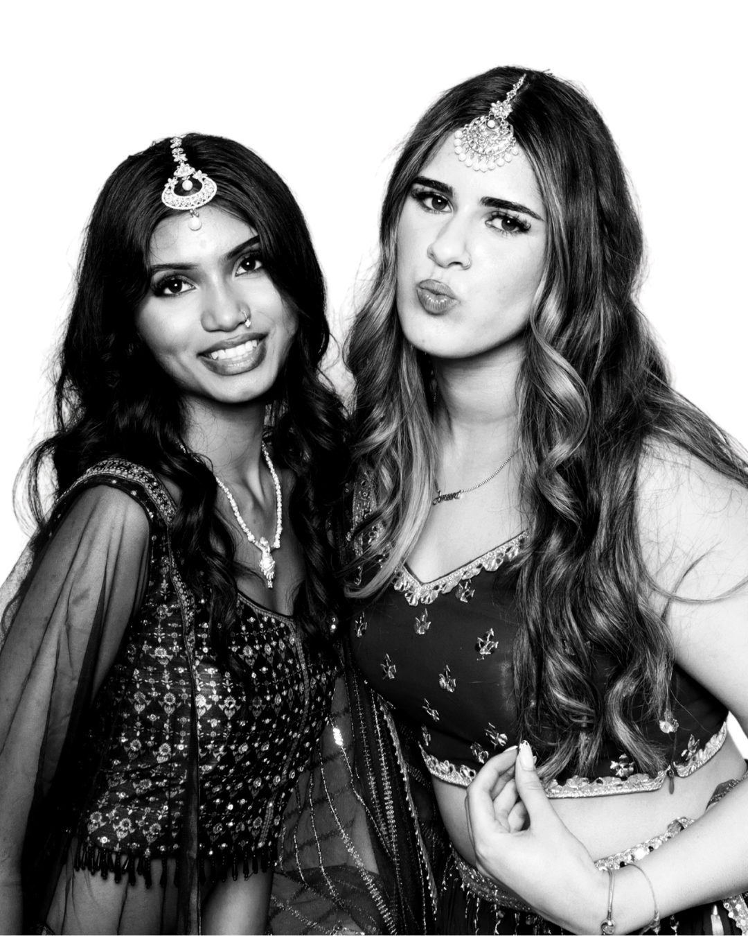Two women wearing traditional Indian attire and jewelry, posing together for a photo, black and white image.