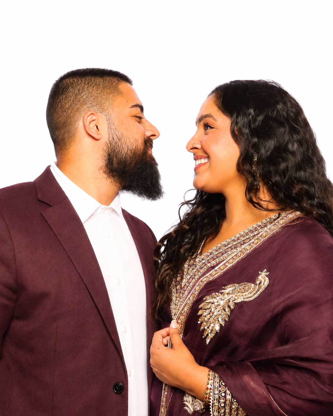 A couple faces each other, smiling with foreheads nearly touching, against a plain white background. The man has a beard and short hair, wearing a burgundy suit and white shirt. The woman has long, curly black hair, wearing traditional maroon and gol