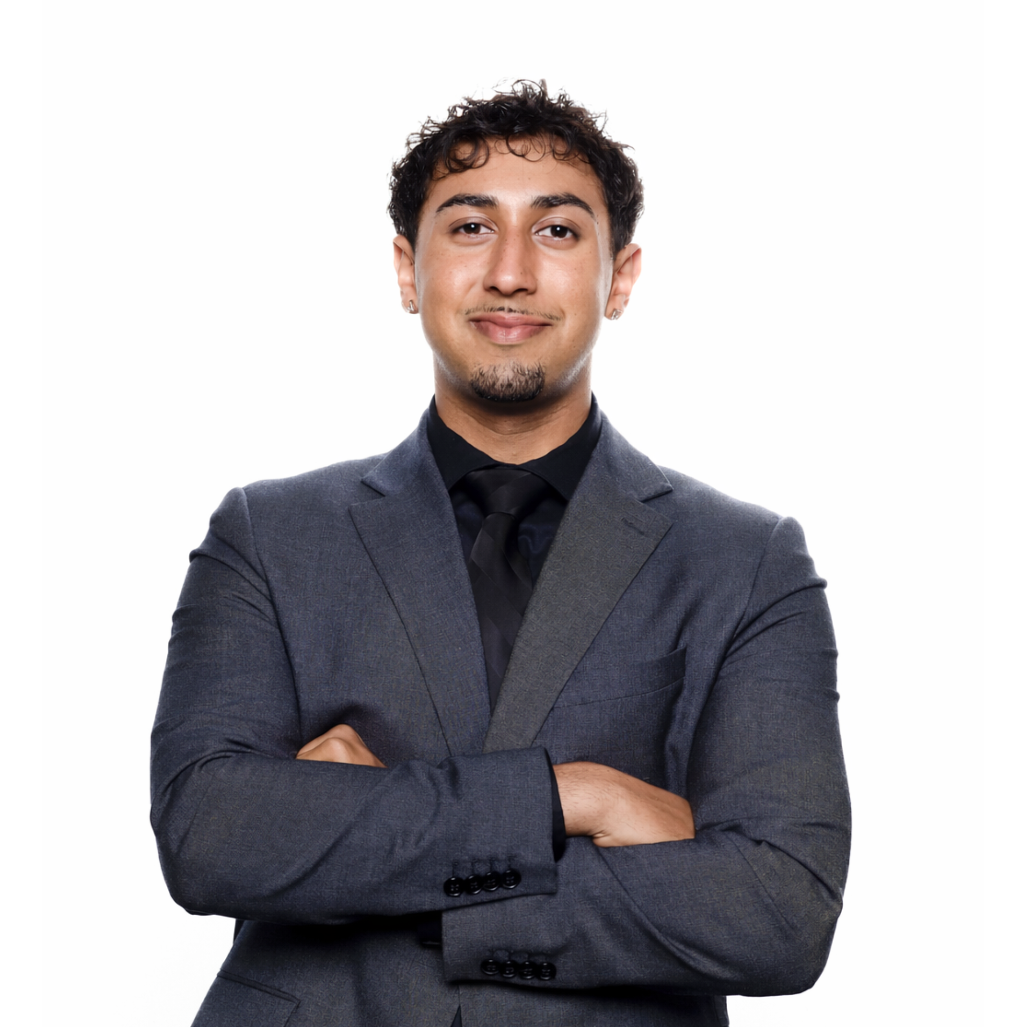 A man with curly dark hair, small earrings, a goatee, wearing a dark gray suit and black shirt, crossing his arms in front of a plain white background.
