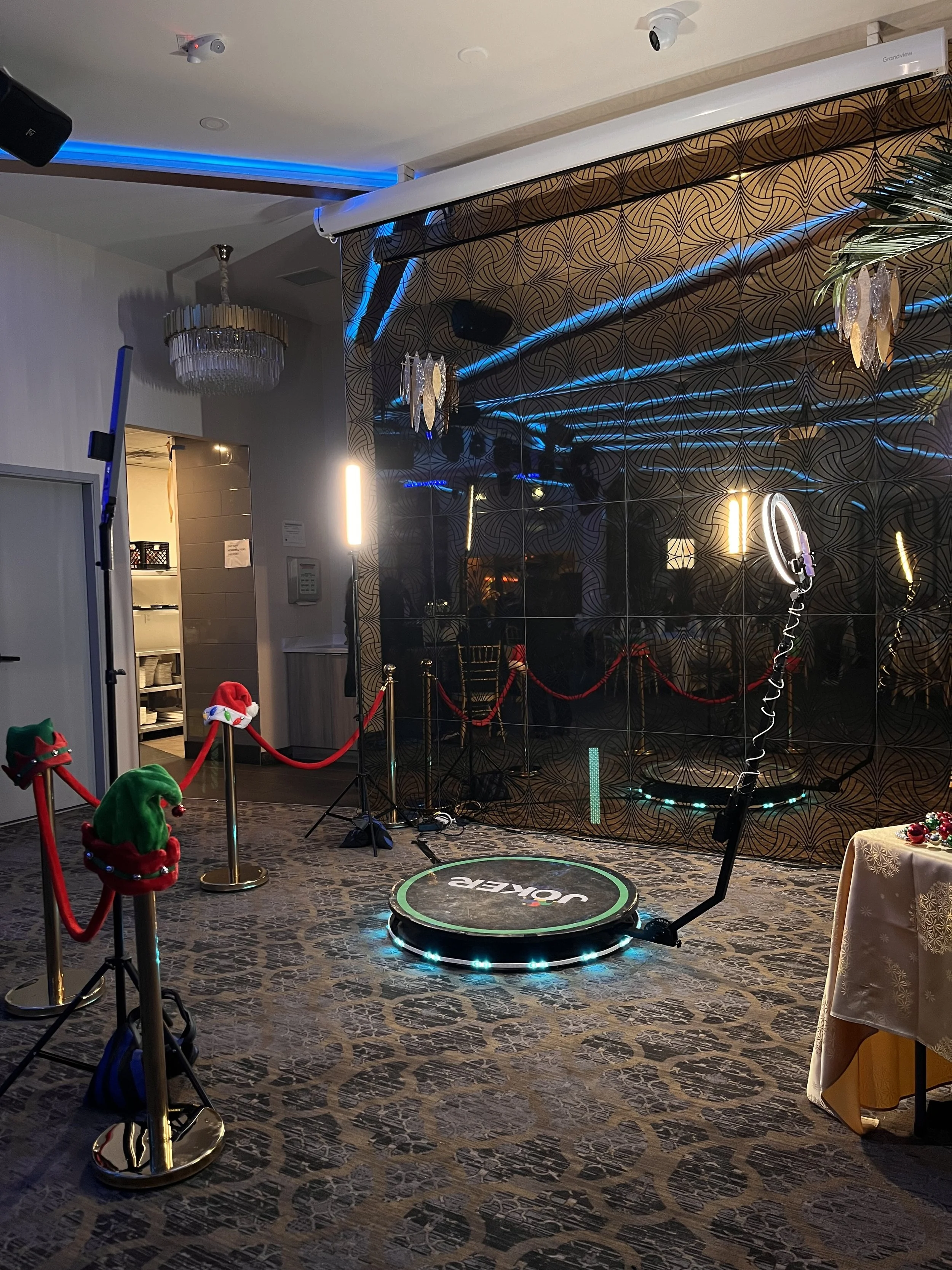 Indoor space decorated for Christmas with a small trampoline, a table with a gold-colored tablecloth, festive ornaments, and Christmas hats on poles. A black reflective wall with blue lighting and decorative hanging lights is in the background. There are security cameras and a spotlight on the ceiling.