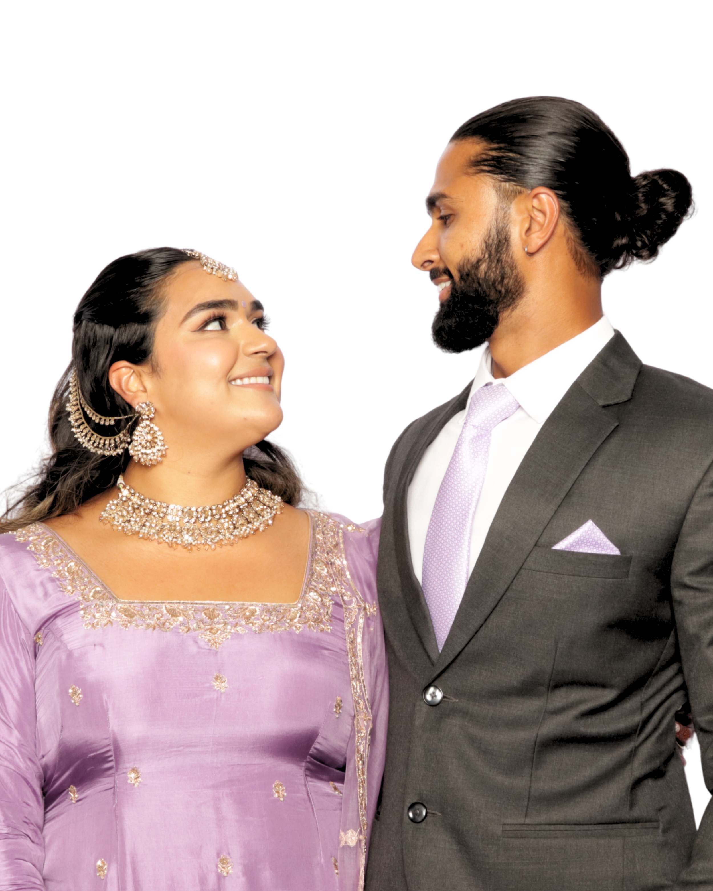 A smiling woman dressed in traditional Indian attire and jewelry, looking up at a man dressed in a gray suit and lavender tie, both with dark hair and beards, against a plain white background.