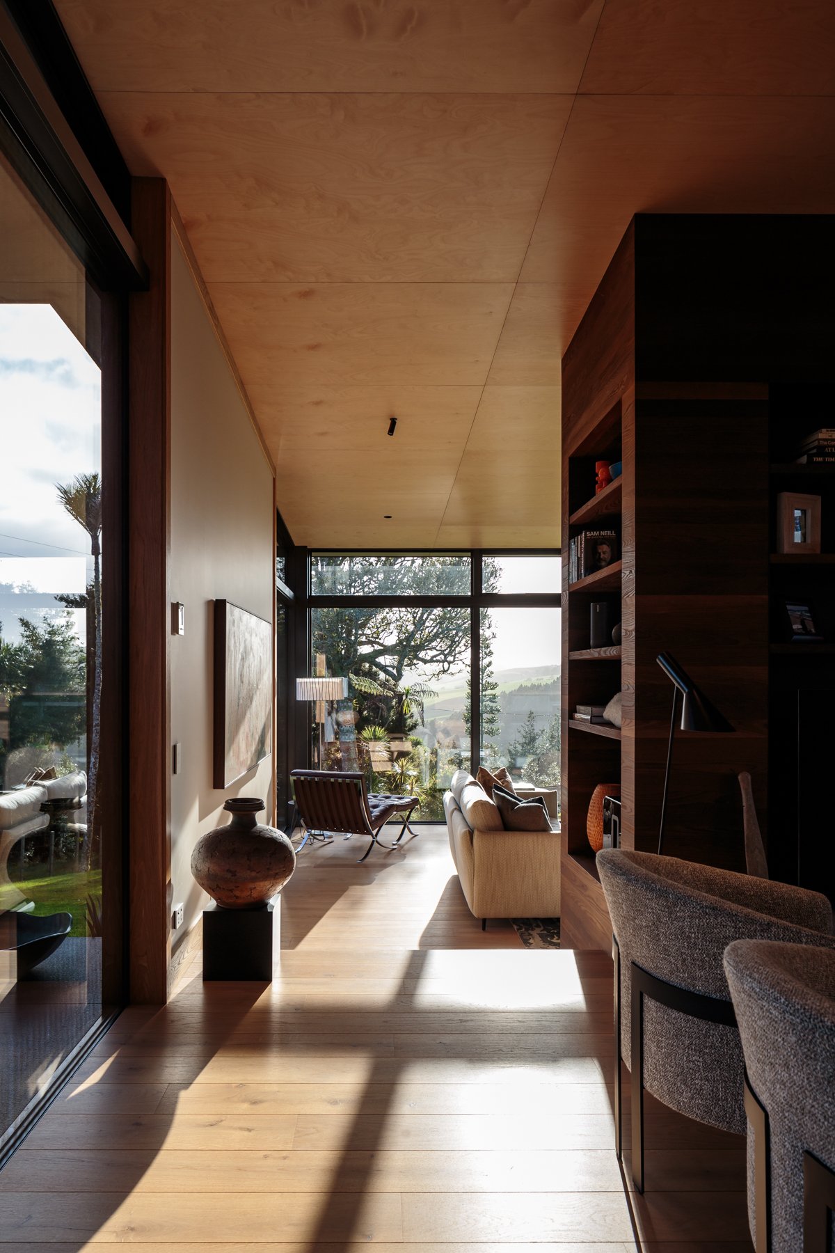 Modern living room with large floor-to-ceiling windows displaying trees and landscape outside, featuring a beige sofa, wooden bookshelves, and decorative vases, with hardwood flooring and wood-paneled ceiling.