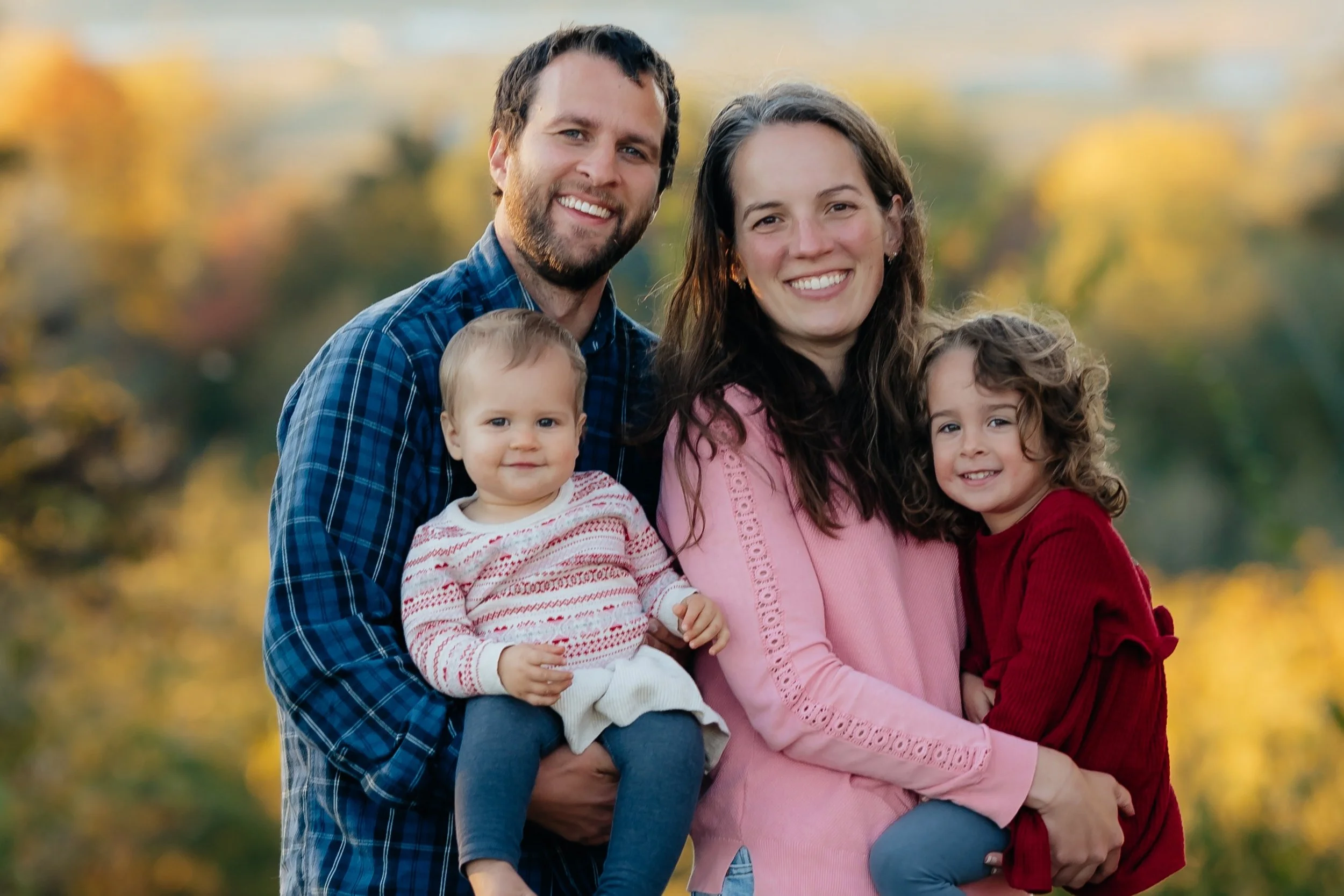 Family of four smiling outdoors during fall, with trees and colorful leaves in the background.
