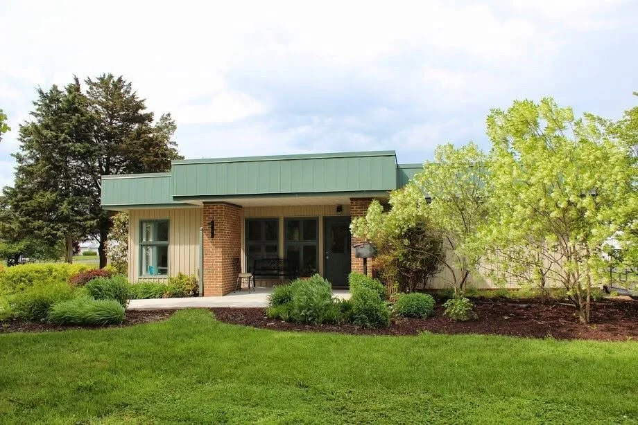 A small house with a green roof, brick and beige exterior walls, a porch with a black bench, surrounded by green grass, shrubs, and trees under a cloudy sky.