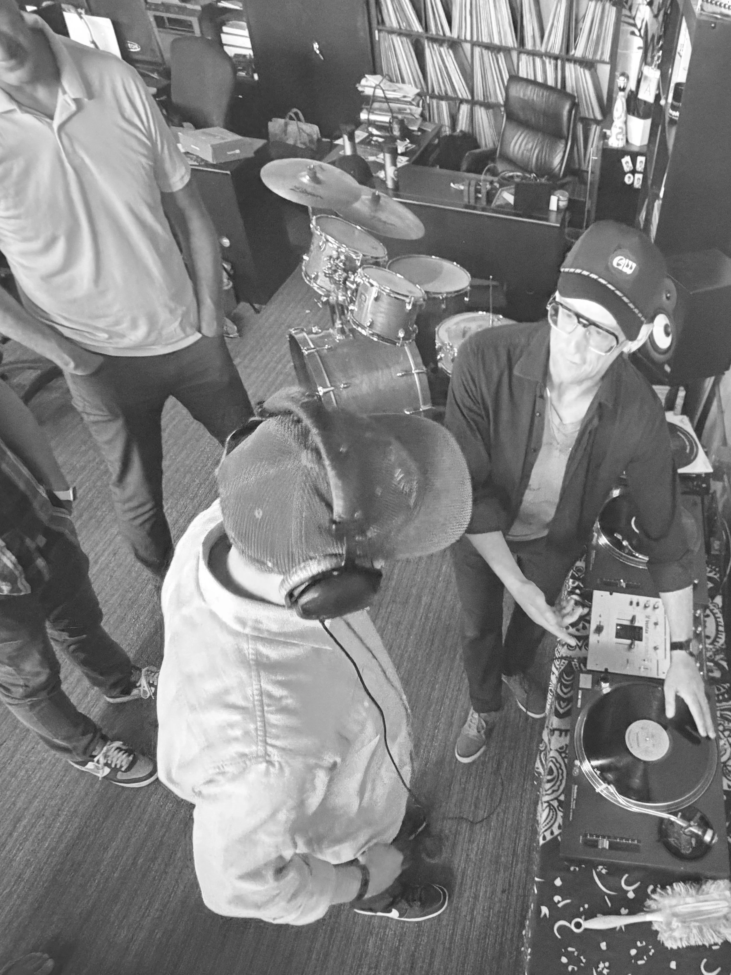 People gathered around a drum set in a room filled with books, a desk, and music equipment, with one person wearing headphones and a cap.