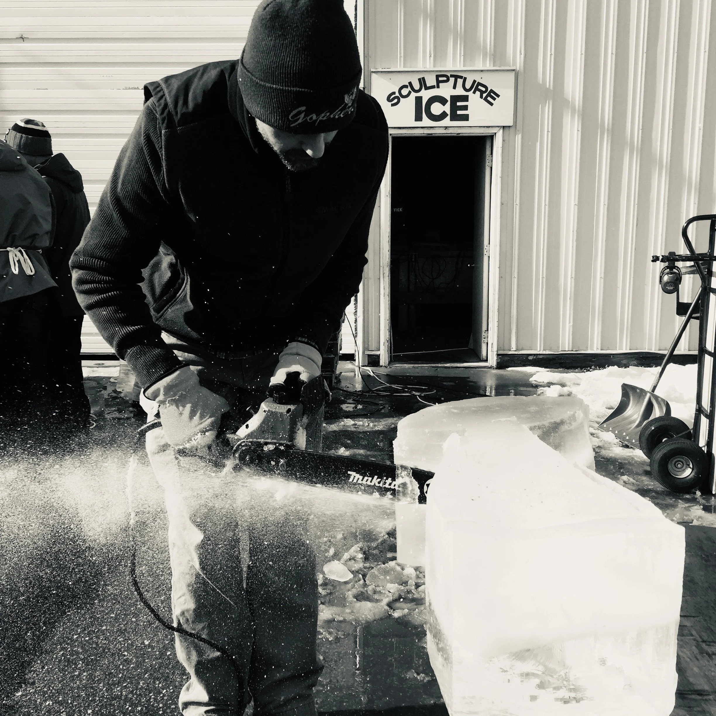 A person in black clothes and a beanie shoveling broken ice on a concrete floor near a building with a sign that reads 'Sculpture Ice'.